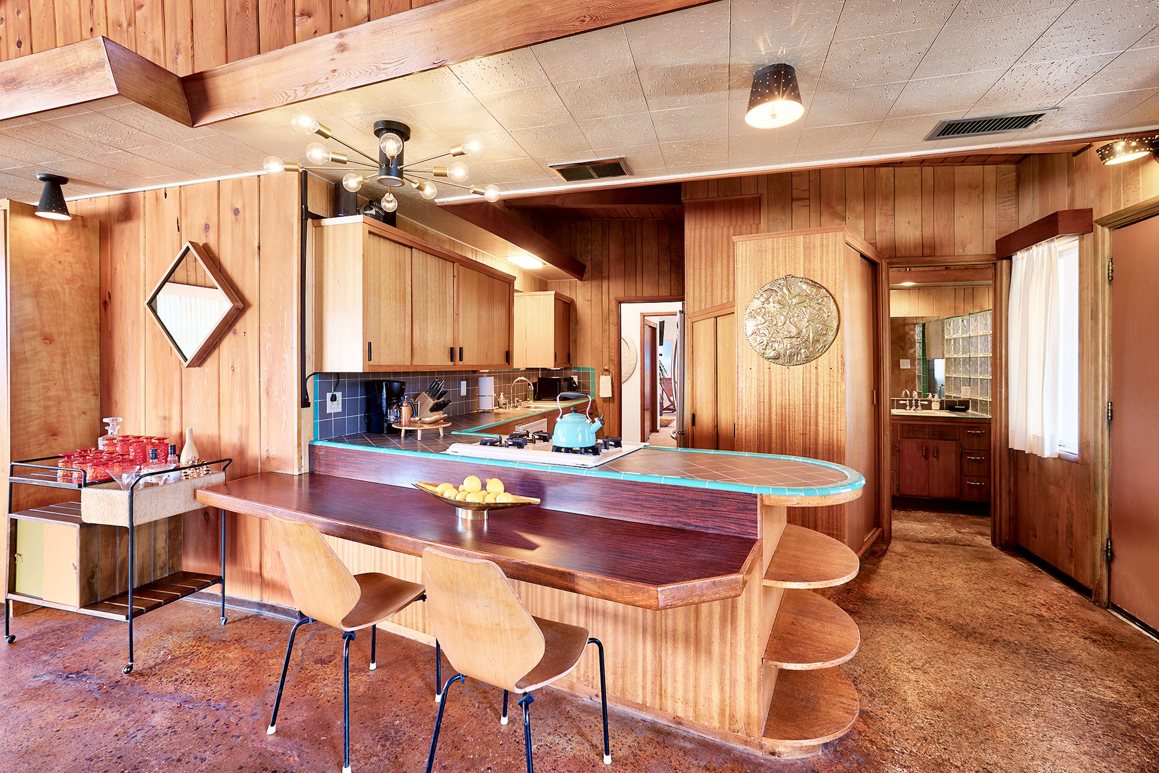 A wooden interior kitchen with a curved countertop, modern light fixtures, and a dining area featuring two chairs and a bowl of lemons.