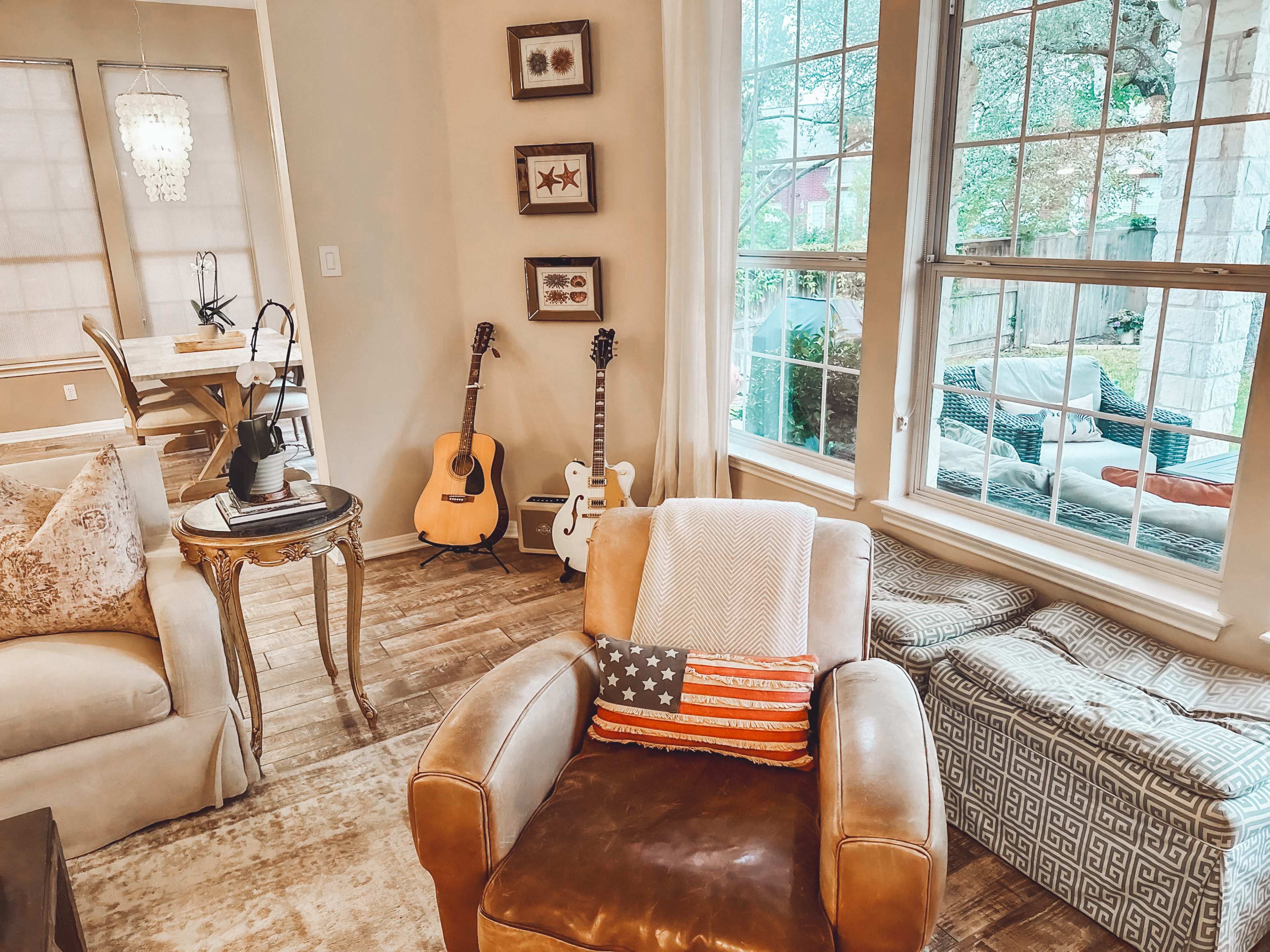 The image shows a cozy living room with a beige armchair, an acoustic guitar on a stand, and large windows letting in natural light.