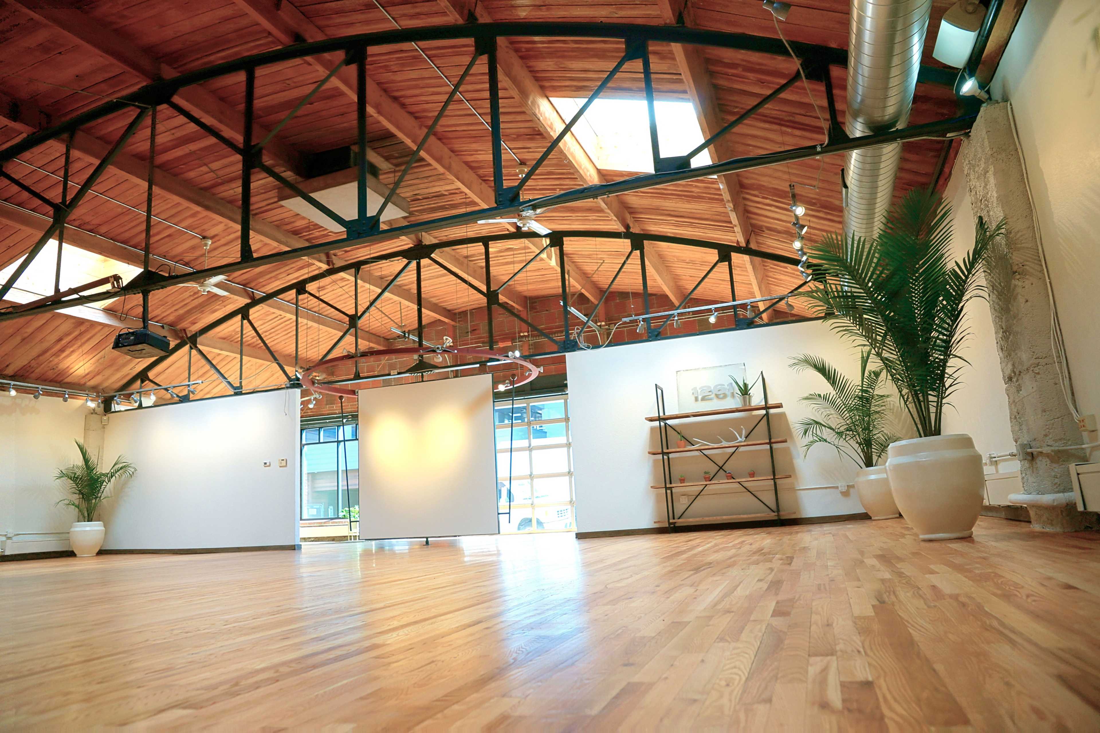 An empty studio space with wooden floors, large windows, and a high wooden-beamed ceiling, featuring potted plants and minimal furnishings.