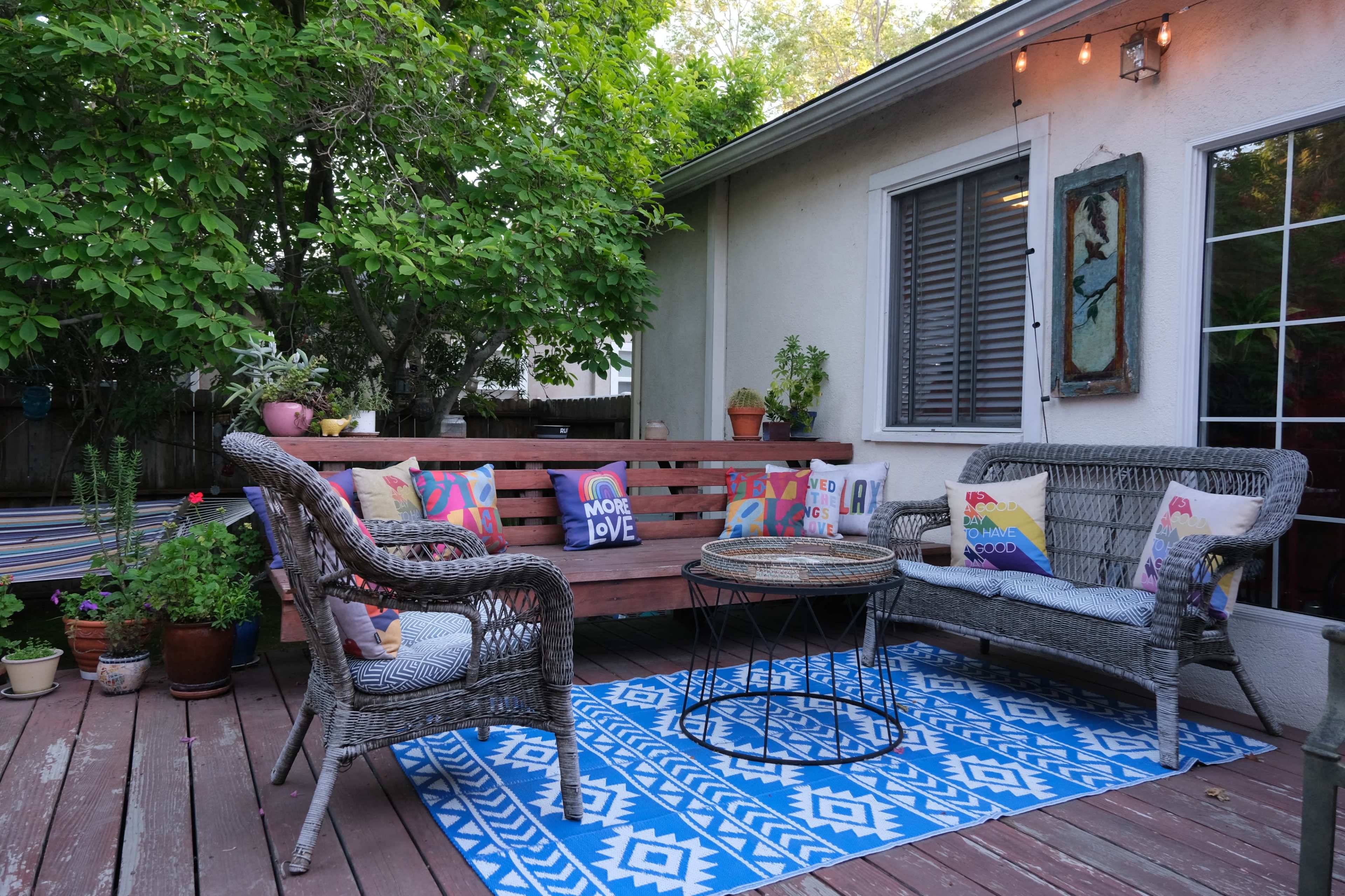 A patio area features a wooden bench with decorative cushions, a circular table, and plants surrounded by greenery.