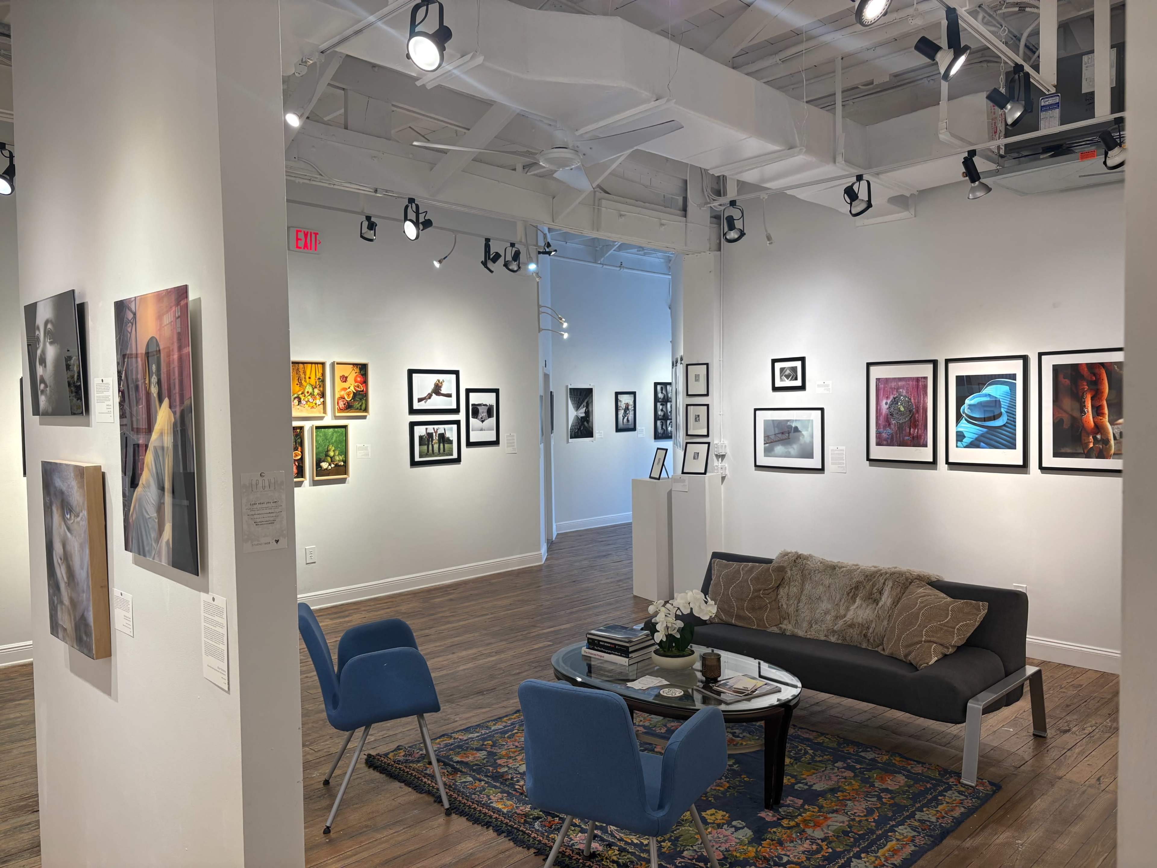 The image shows a contemporary art gallery with various framed artworks displayed on white walls, and a seating area featuring a sofa and blue chairs.