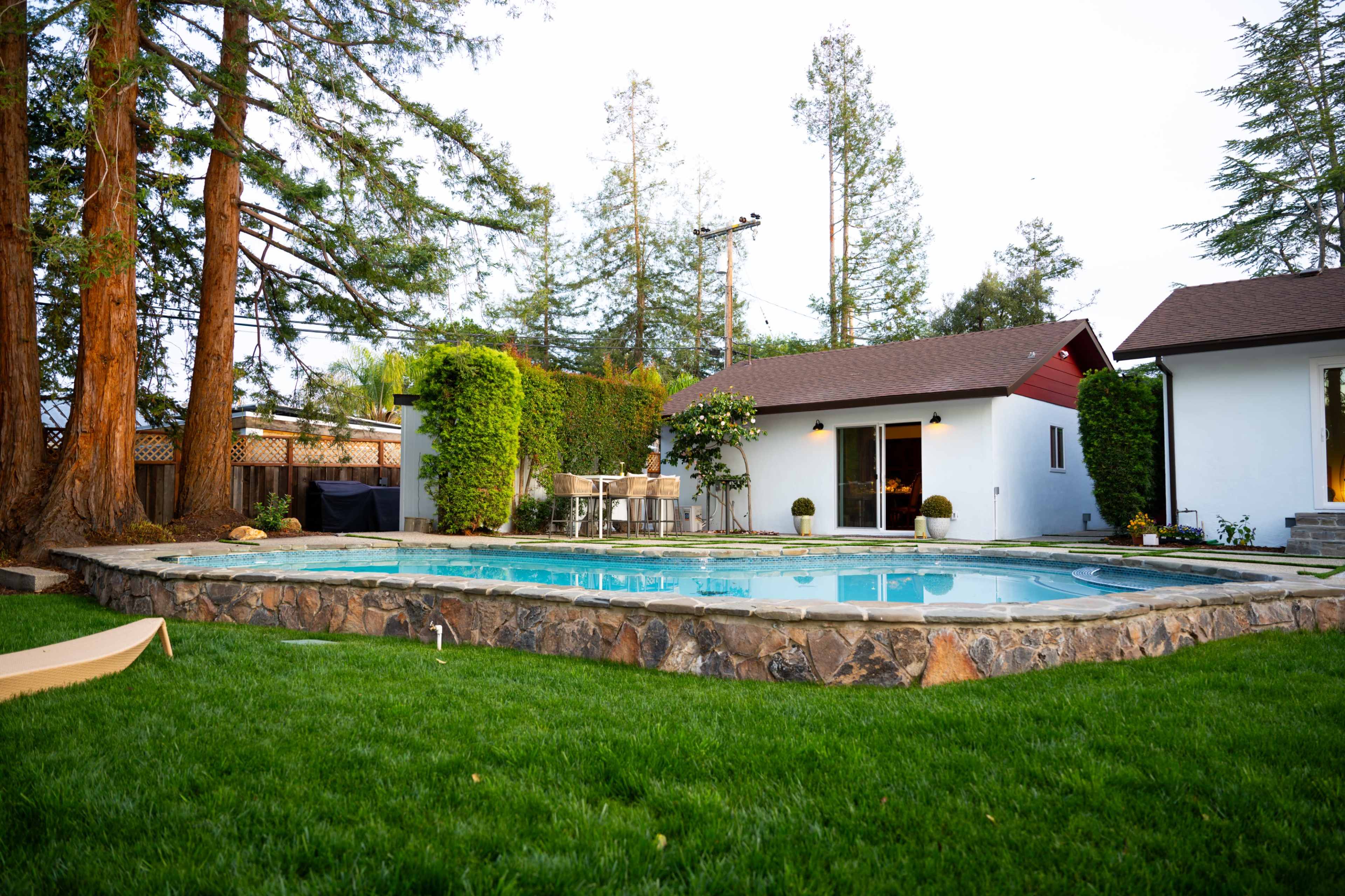 The image shows a backyard featuring a swimming pool surrounded by a stone patio, lush green grass, and two adjacent buildings.
