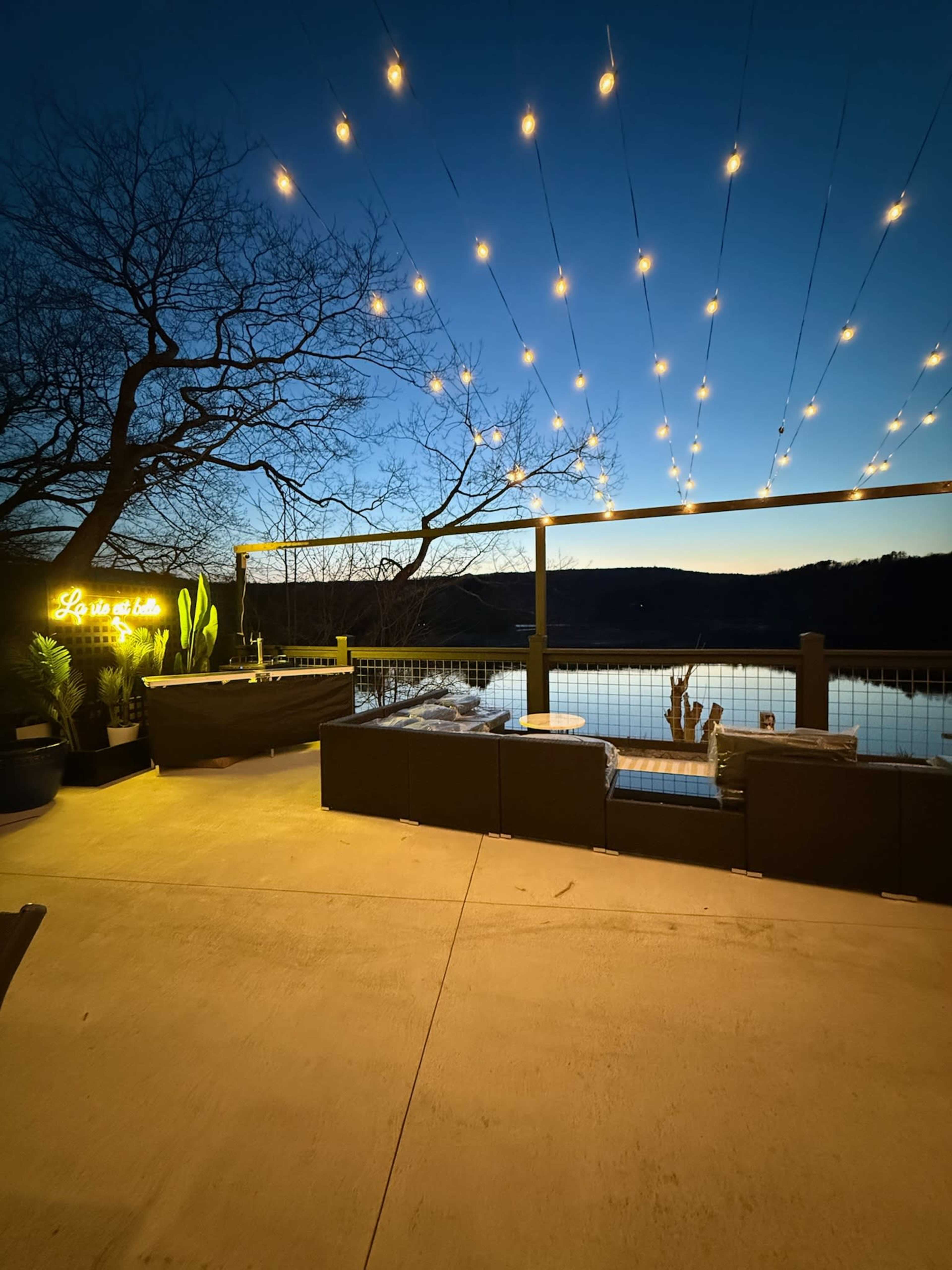 The scene features a patio area with string lights overhead, overlooking a body of water at dusk, with seating and decorative plants arranged around the space.