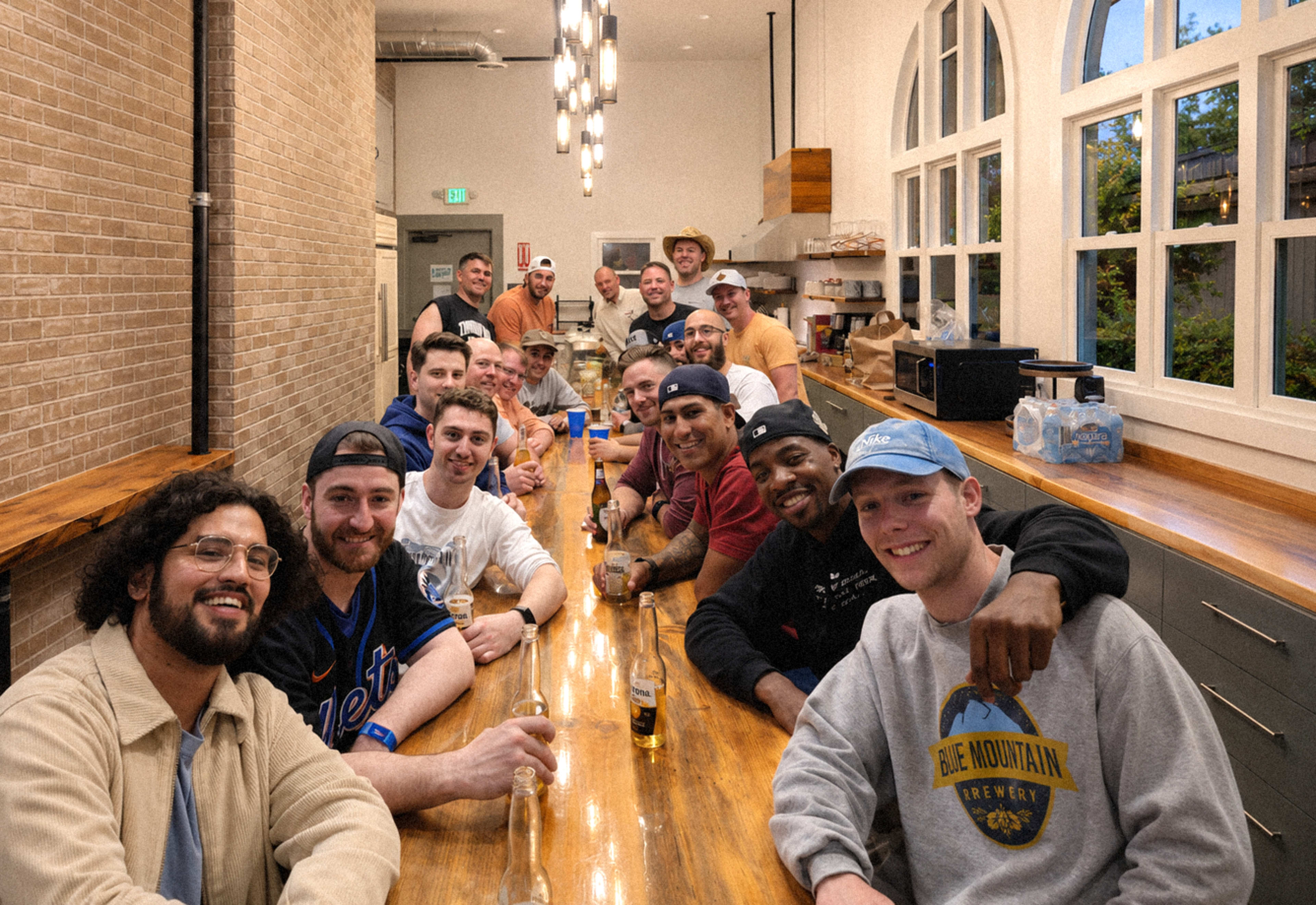 A group of twenty men sits together at a long wooden bar in a well-lit room, smiling and posing for the camera.