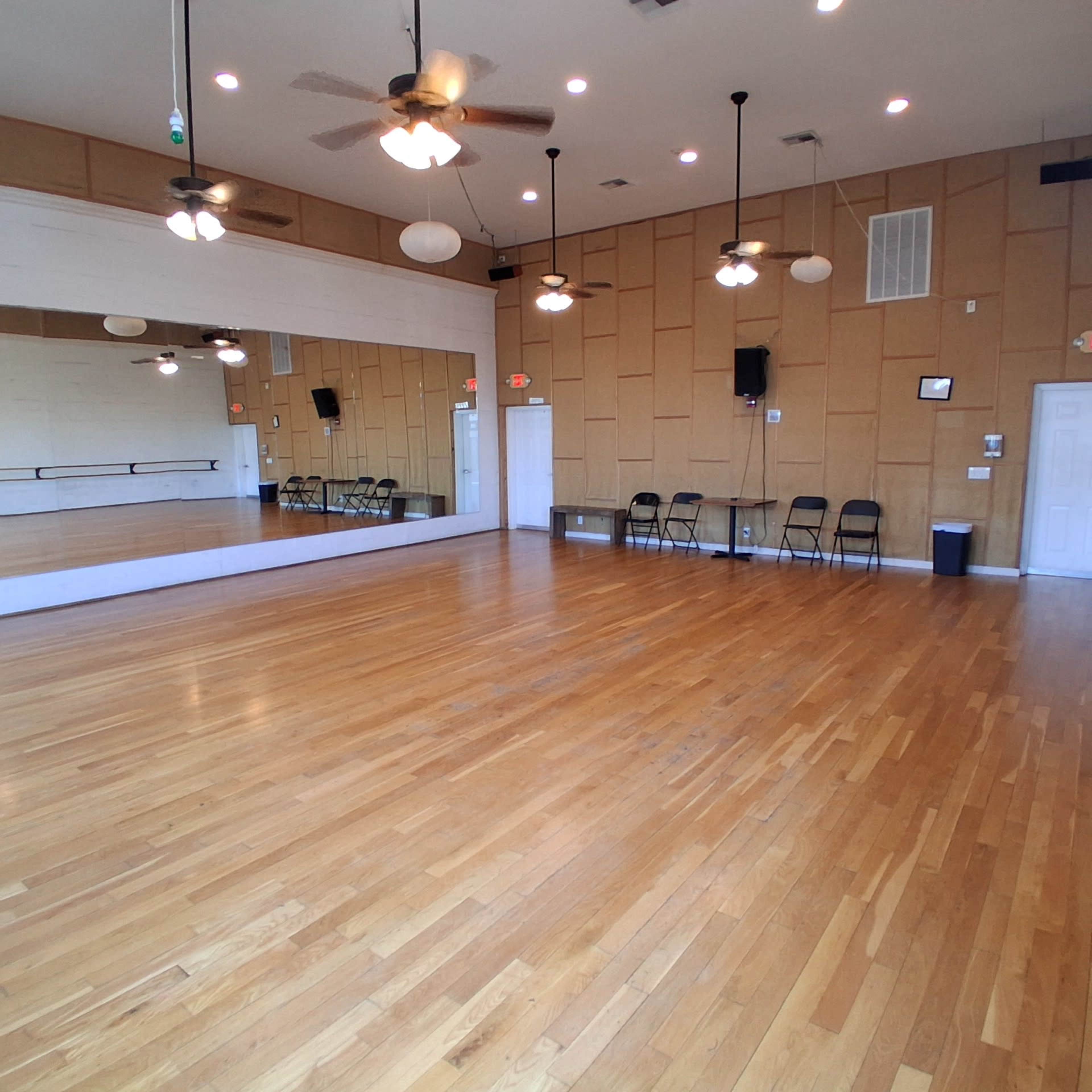 The image shows an empty studio space with wooden floors, wall mirrors, and several folding chairs arranged against the wall.