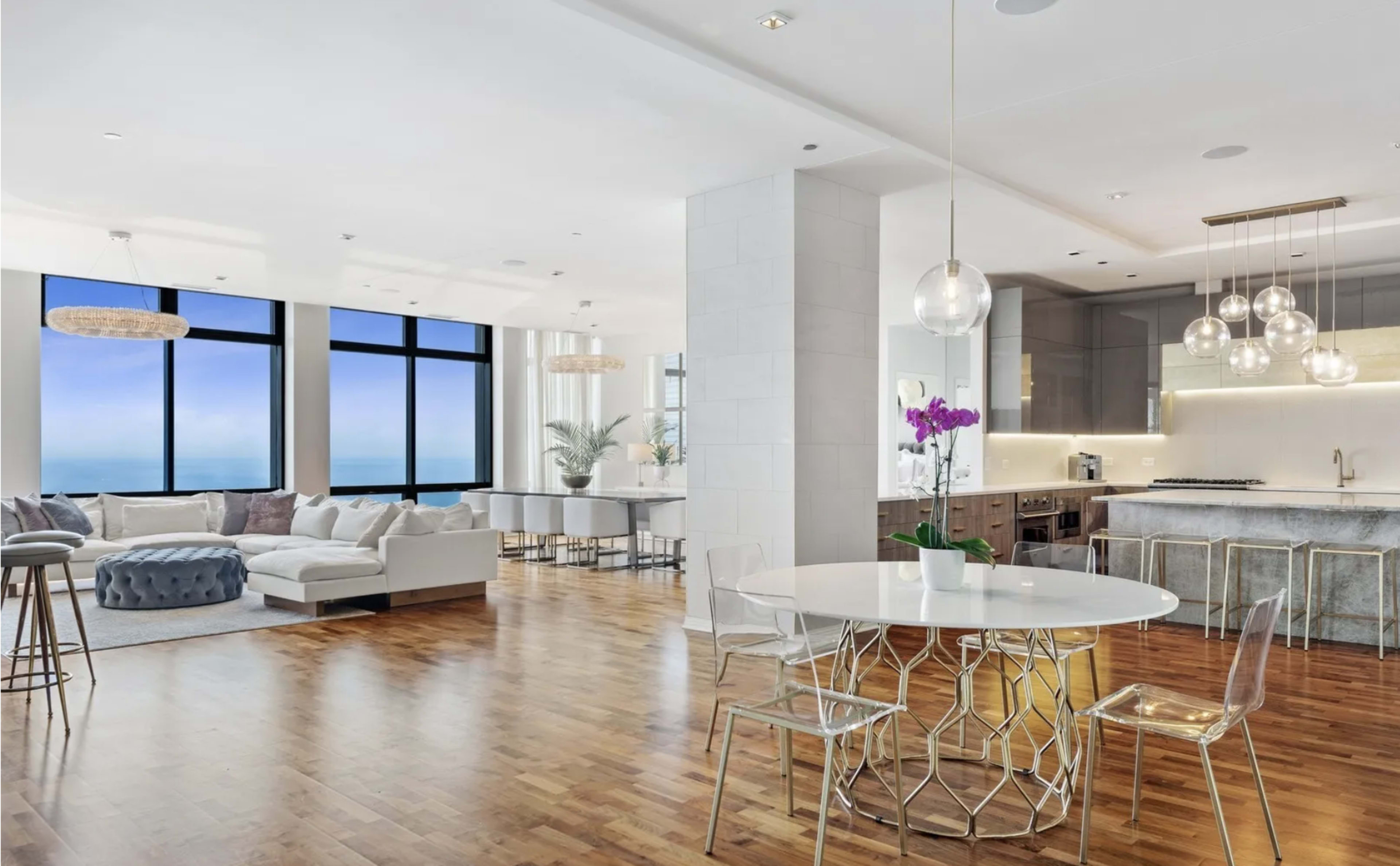 The image shows a spacious modern apartment with large windows overlooking the ocean, featuring a living area, dining space, and a sleek kitchen.