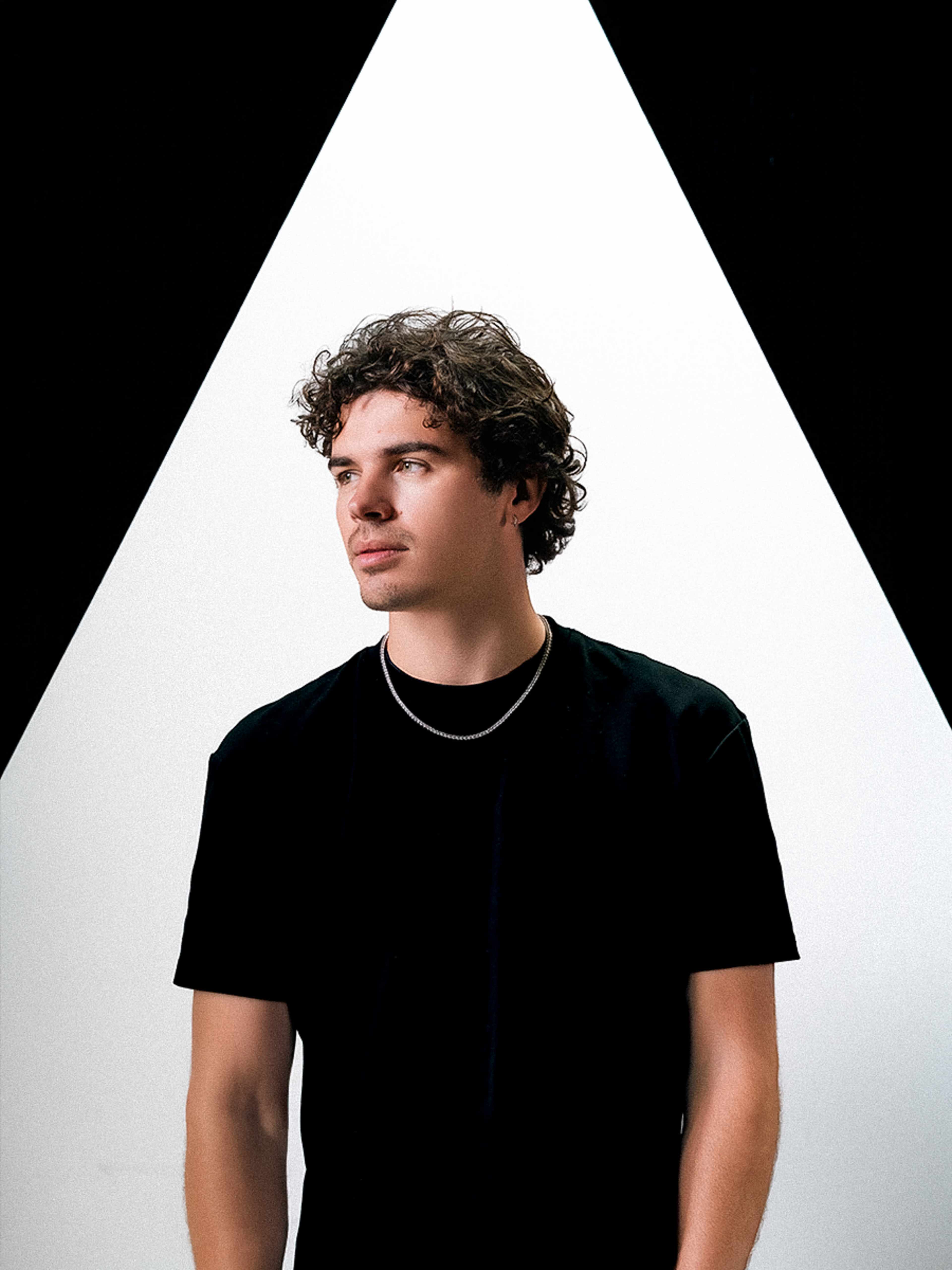 A person with curly hair is standing in front of a stark white triangle illuminated against a black background.