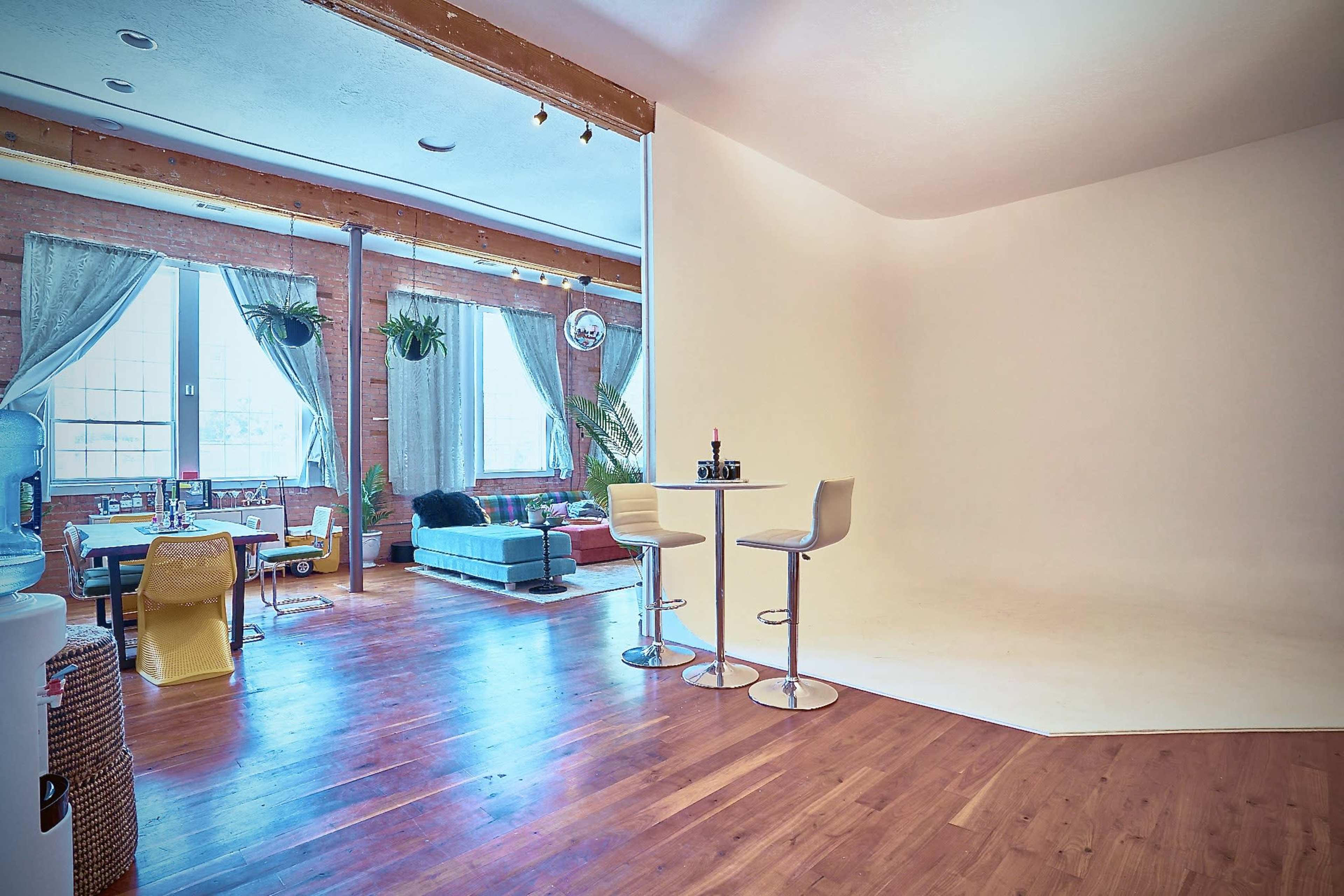 A spacious interior features large windows, wooden floors, a cozy seating area, and a white backdrop for photography.