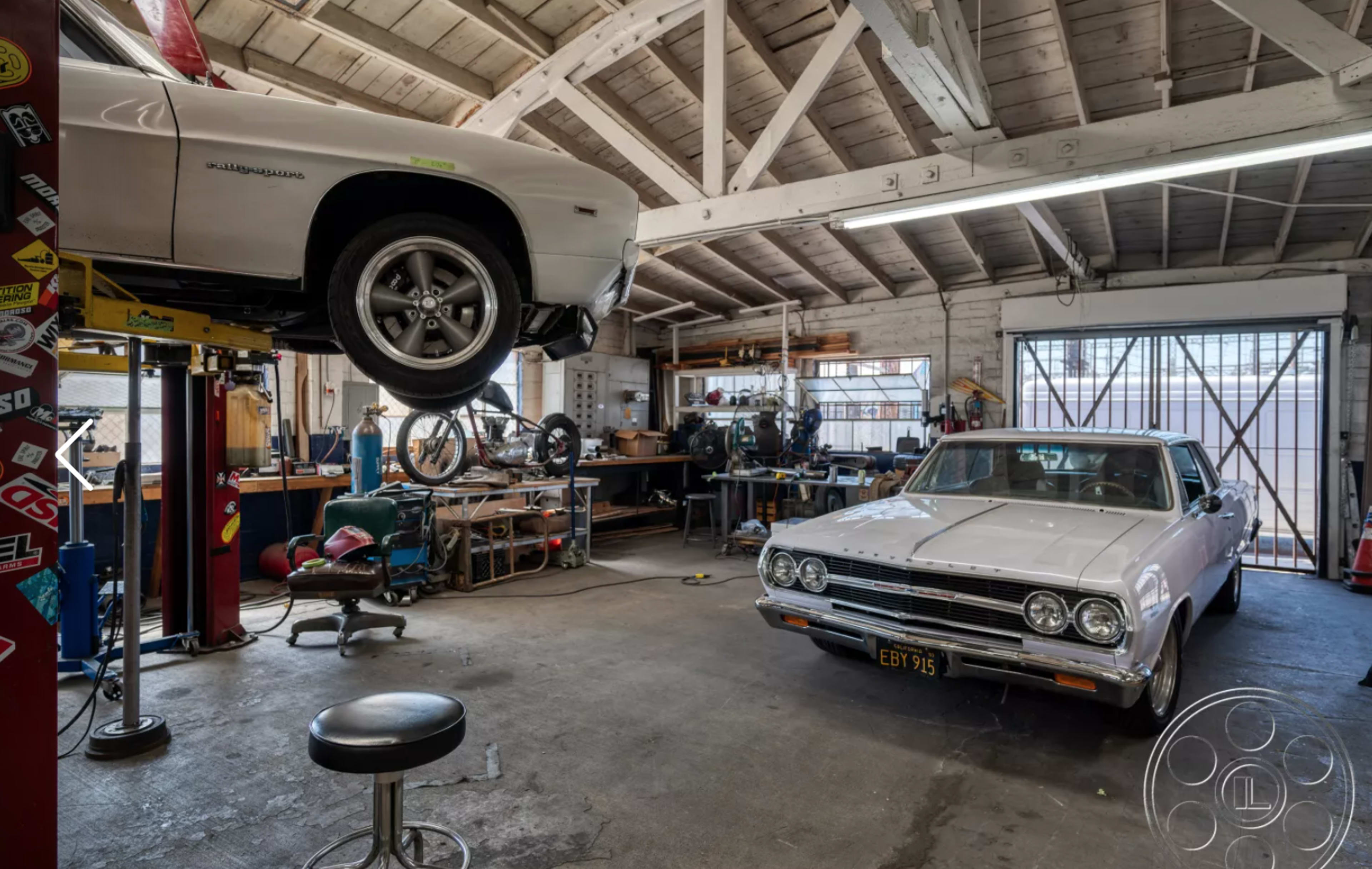 A well-equipped garage features two classic cars, one on a lift and the other parked, surrounded by tools and workbenches.