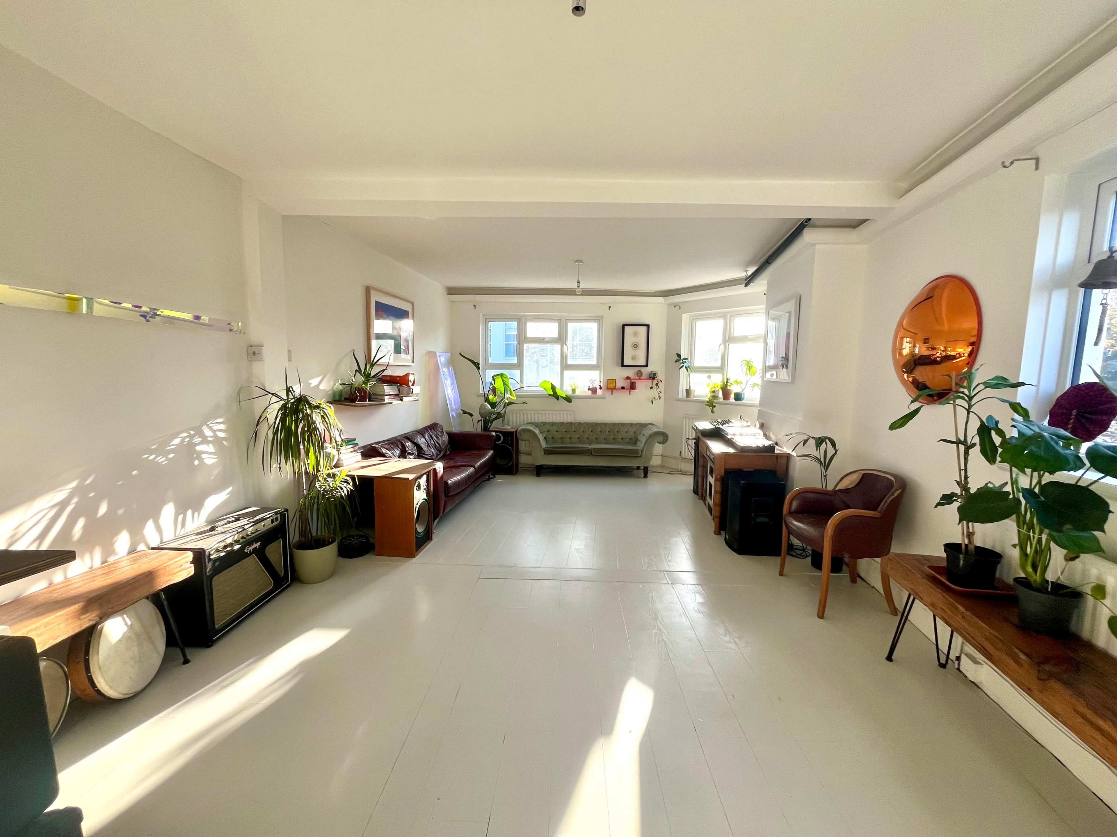 The image shows a bright, open living space featuring a mix of seating areas, plants, and natural light coming through the windows.