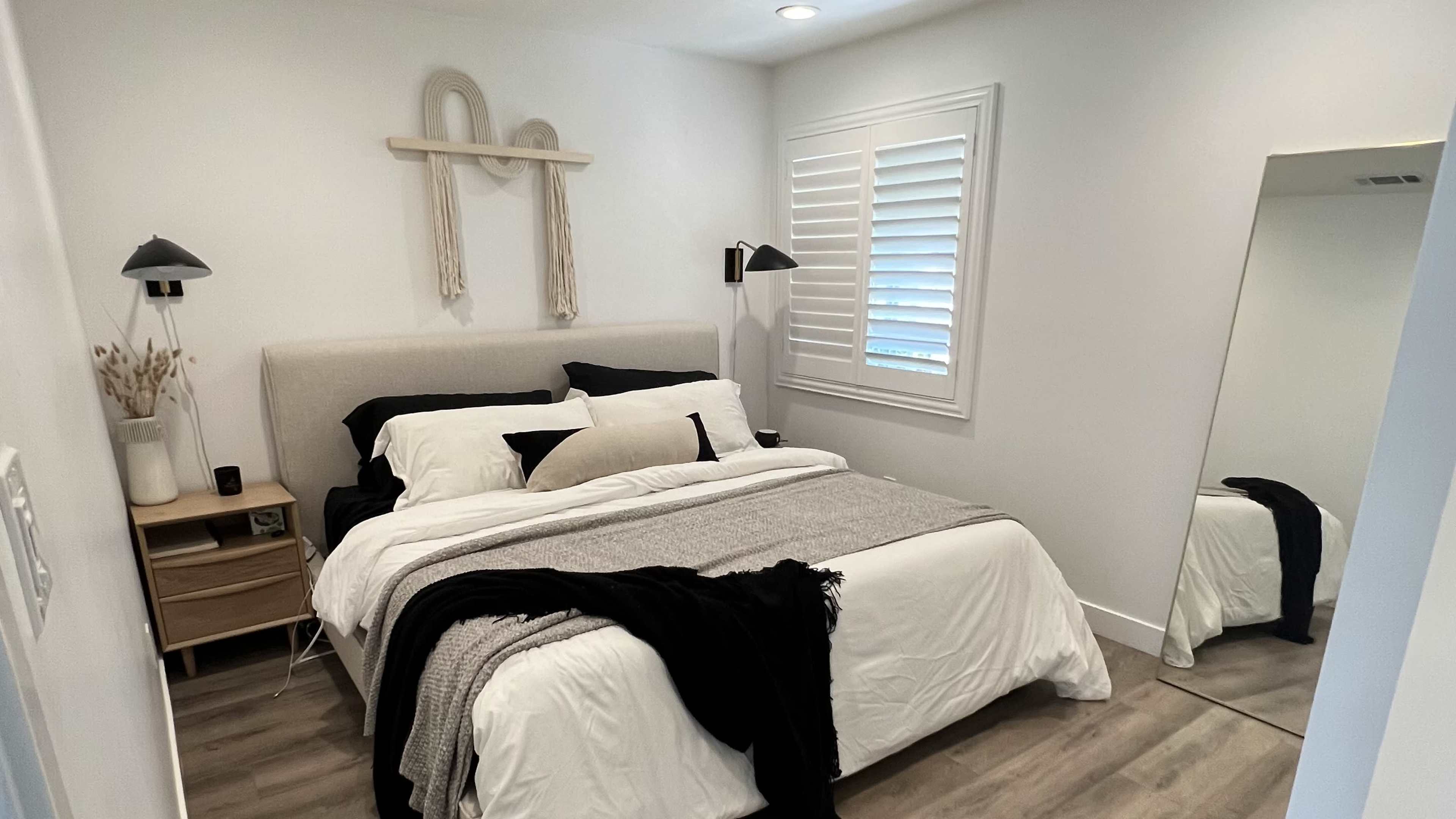 A modern bedroom with a bed adorned in white linens and a gray throw, accompanied by bedside tables and a floor mirror against white walls.