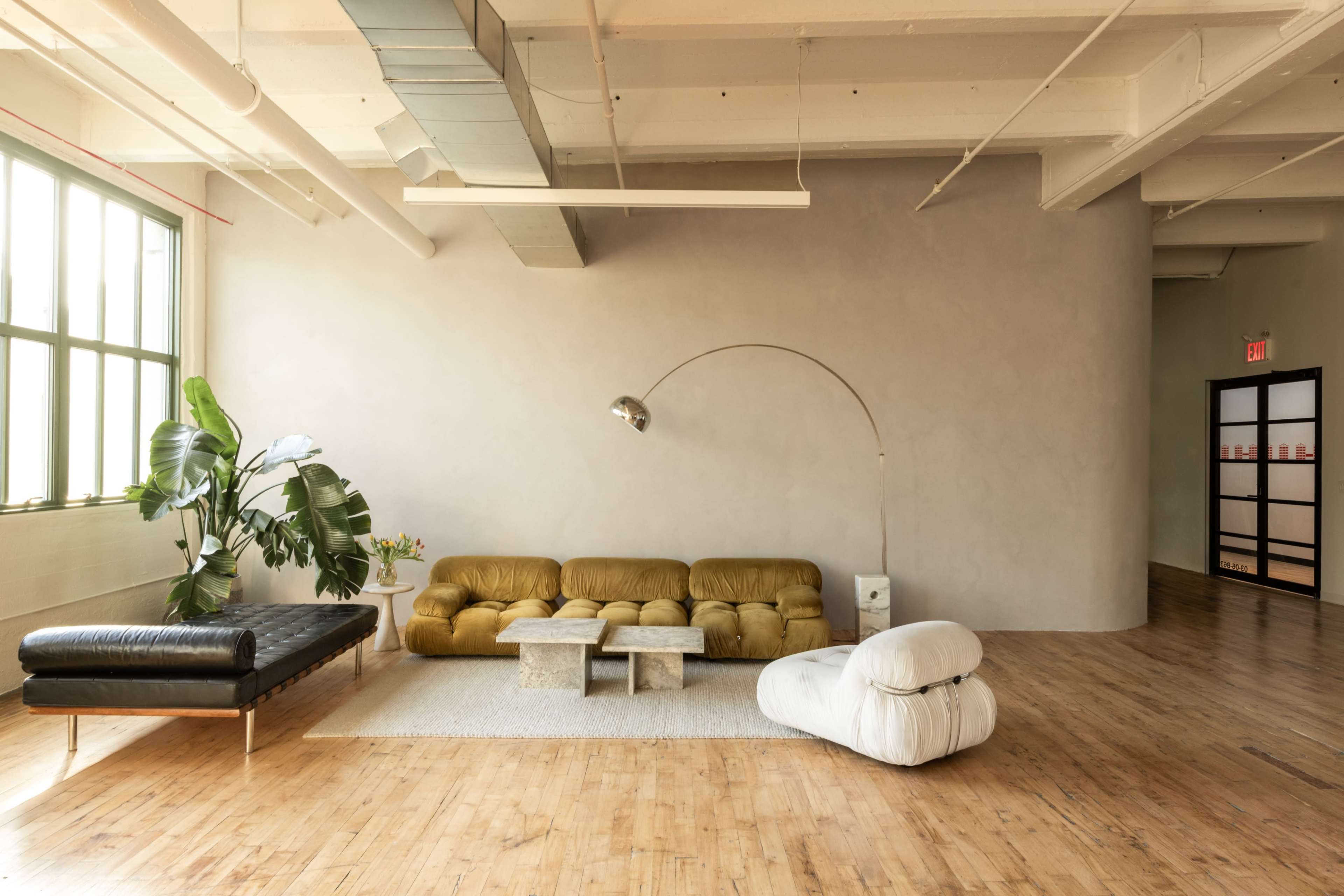 The image shows a spacious, modern interior with a yellow tufted sofa, a black leather couch, a white bean bag chair, and a large potted plant, all set on a wooden floor.