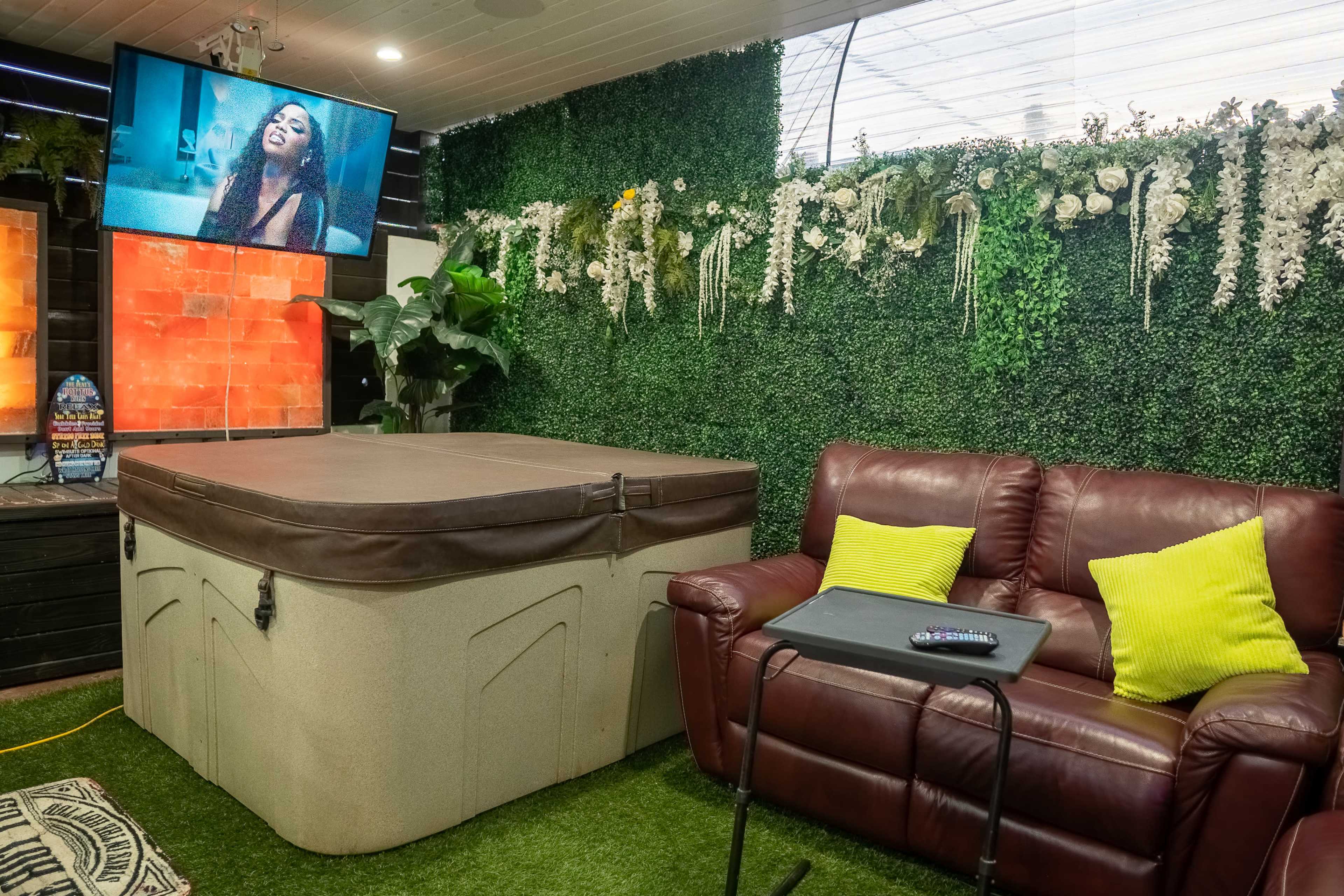 A cozy indoor space with a hot tub, a leather couch, a side table, and a television displaying a woman on screen, surrounded by green plants and decorative wall features.