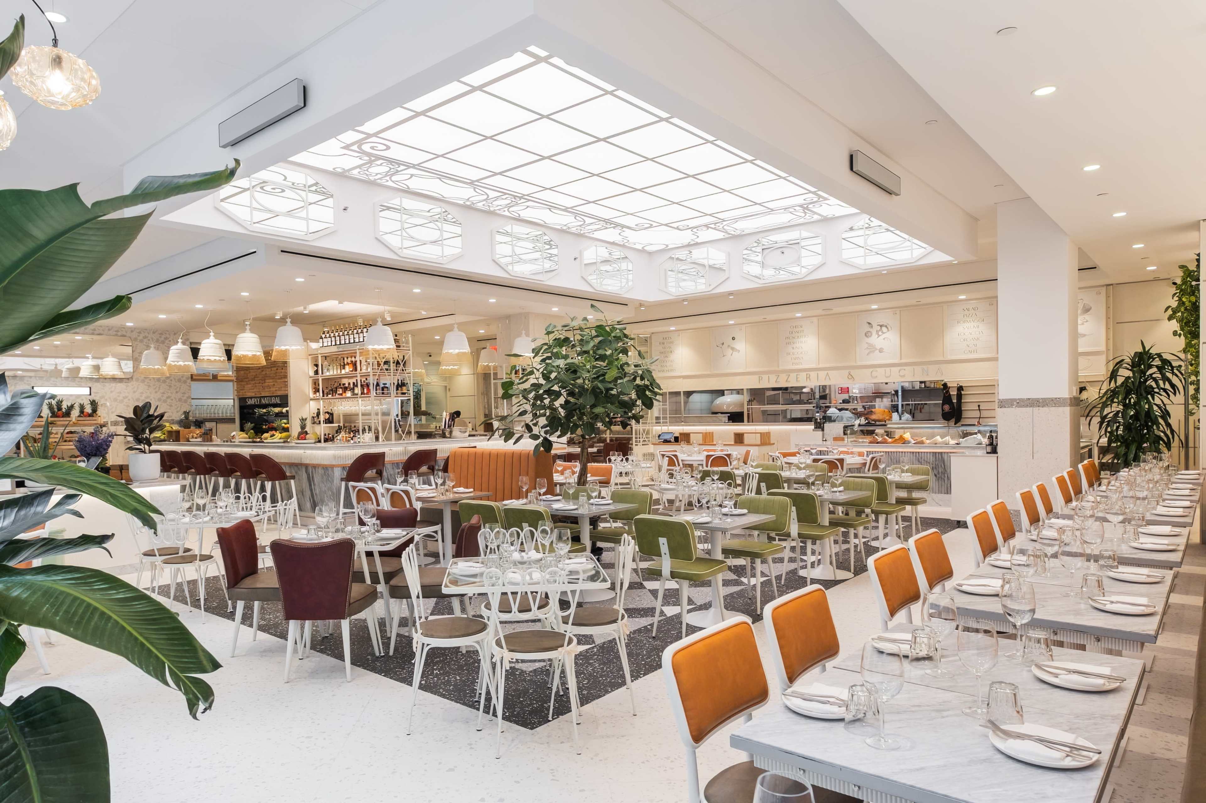 The image shows a spacious, modern restaurant with a mix of green, orange, and white furniture, large windows, and a well-lit open kitchen.