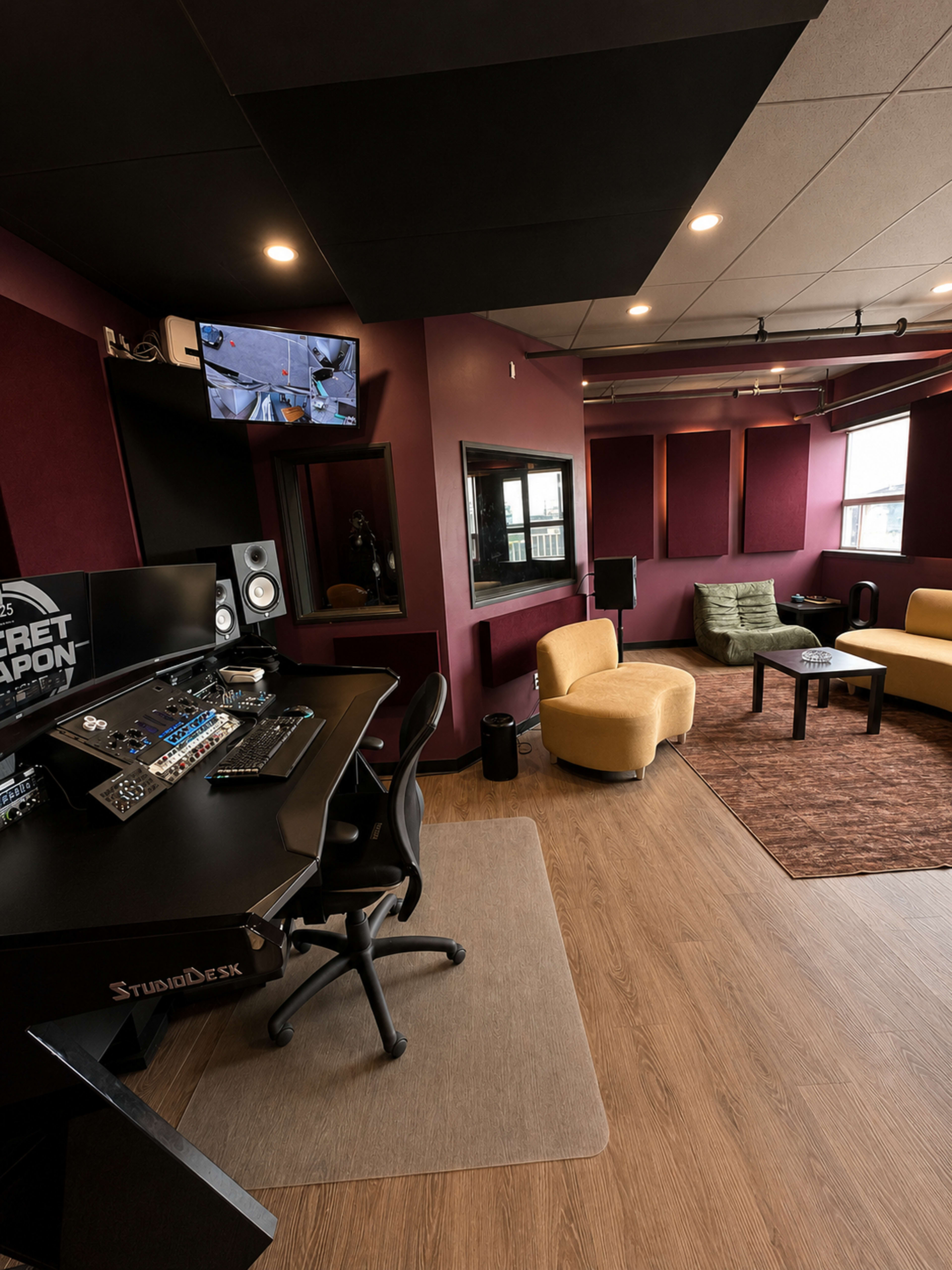 The image shows a modern audio recording studio with a black mixing desk, comfortable seating, and soundproofing panels on the walls.