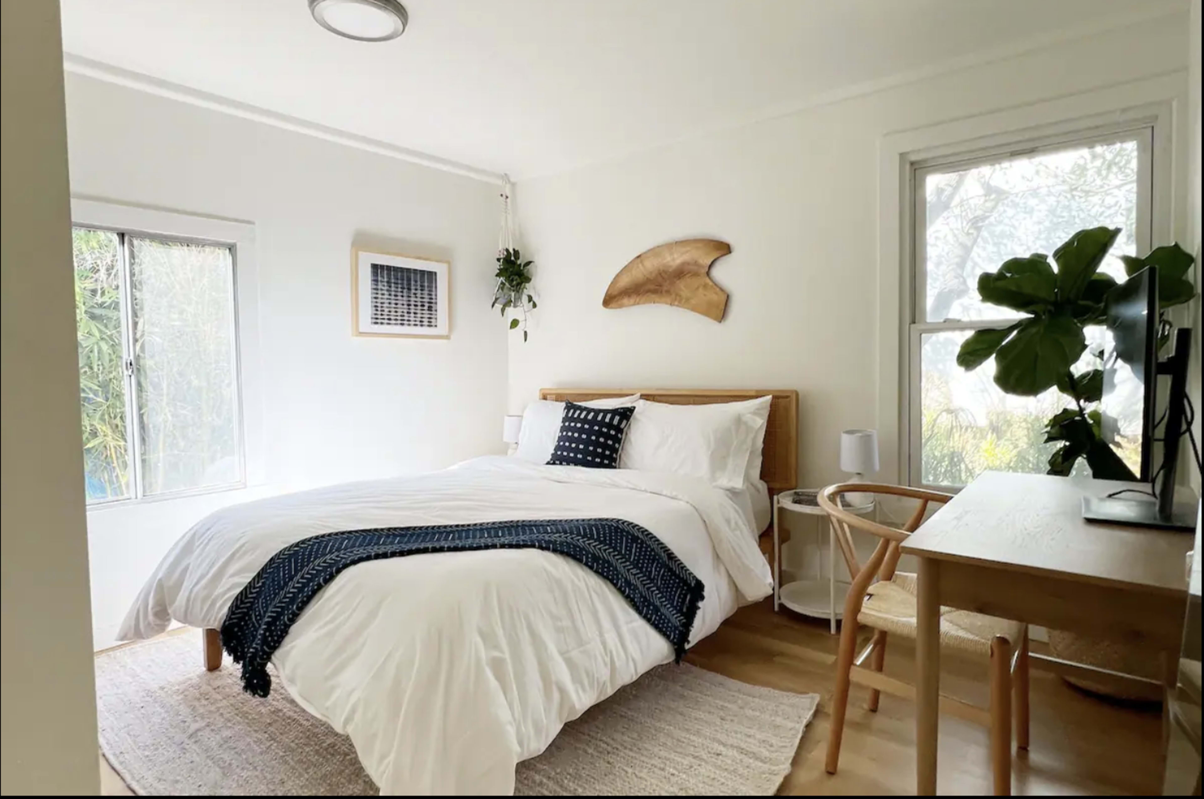 A bright bedroom featuring a bed with white bedding, a wooden headboard, and a desk beside a window.