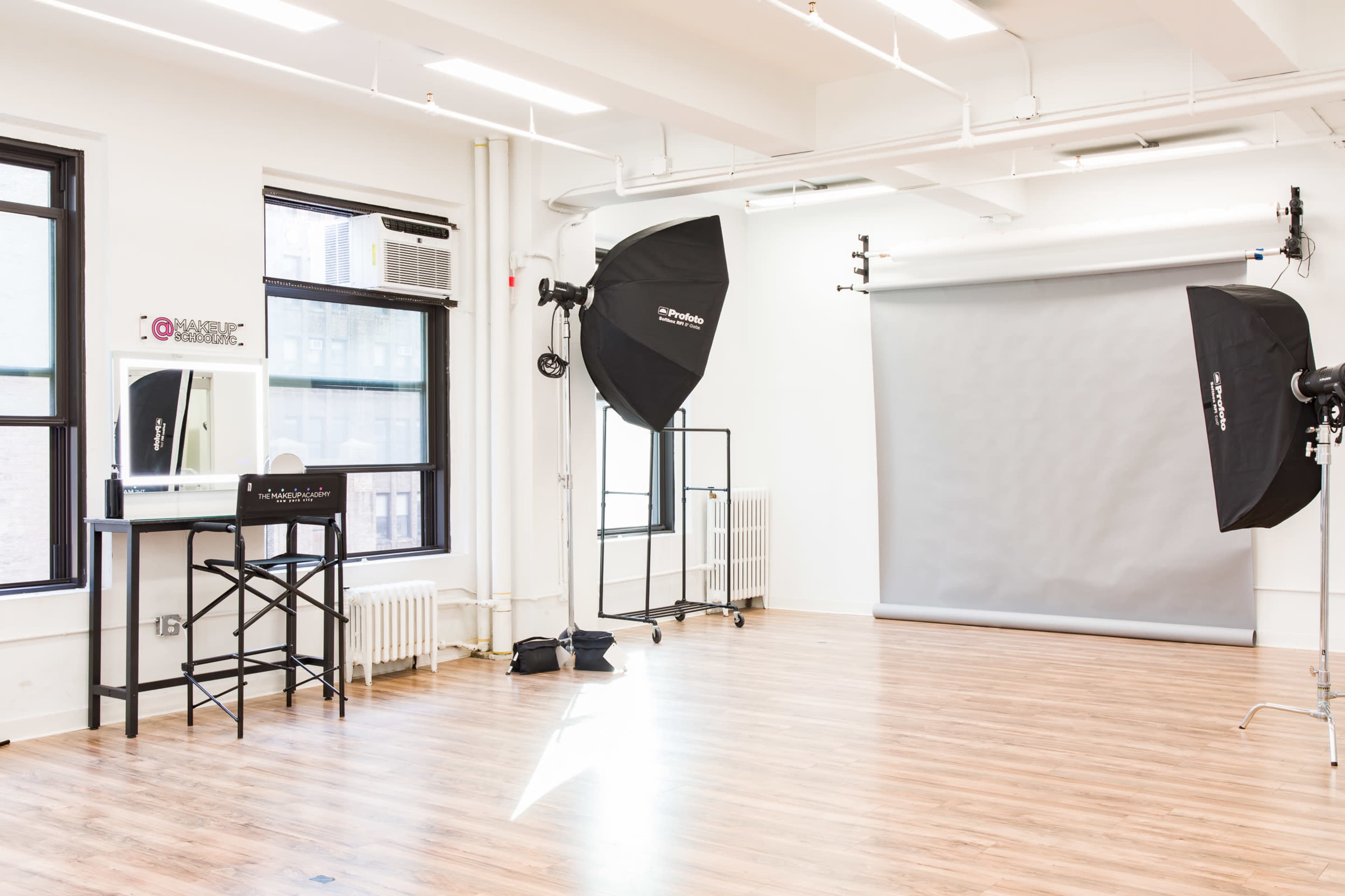 A photography studio with a large gray backdrop, two softboxes set up for lighting, and a makeup station with a mirror and chair.
