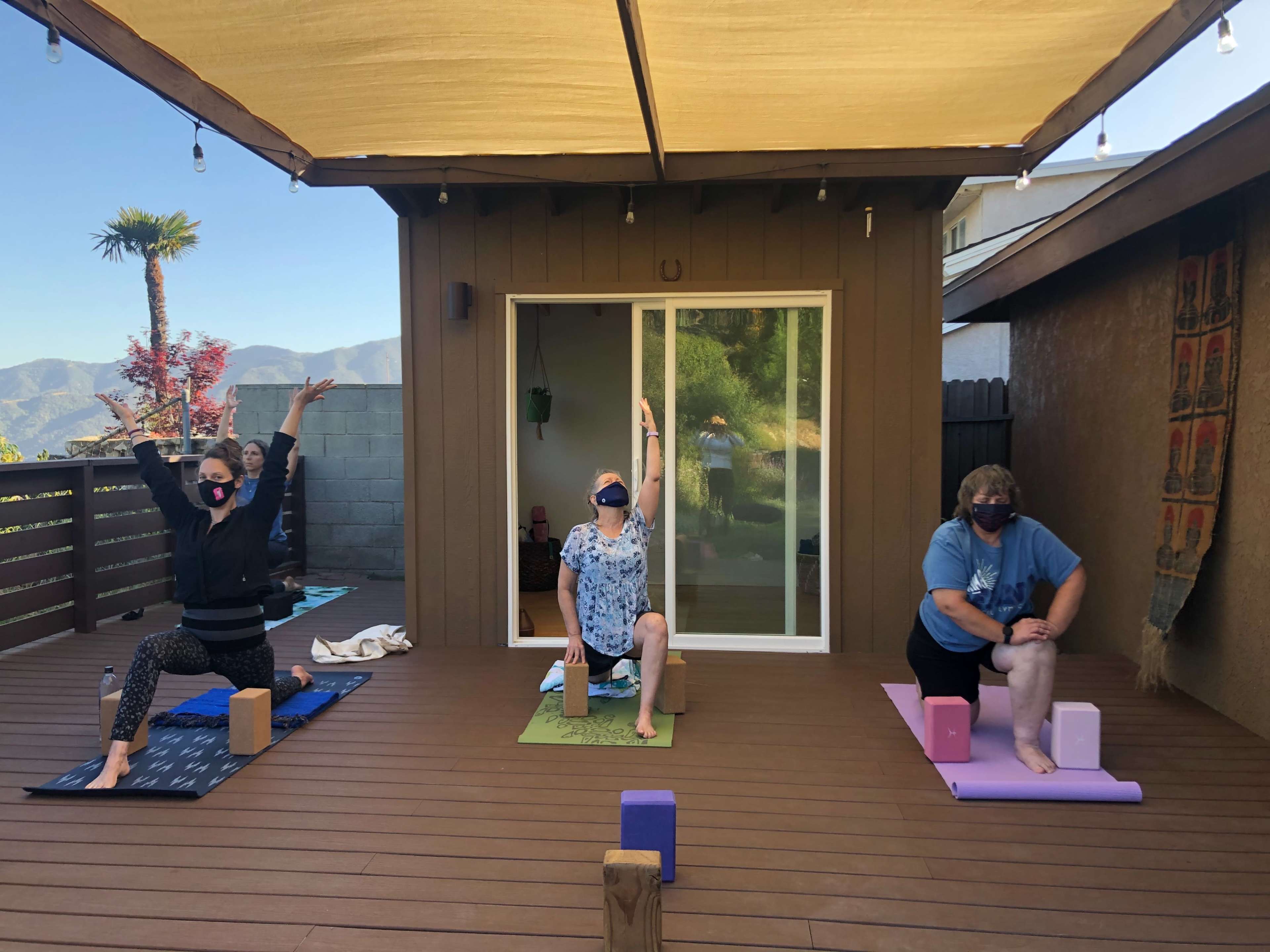 Three individuals practice yoga on a wooden deck, with a mountain view in the background.