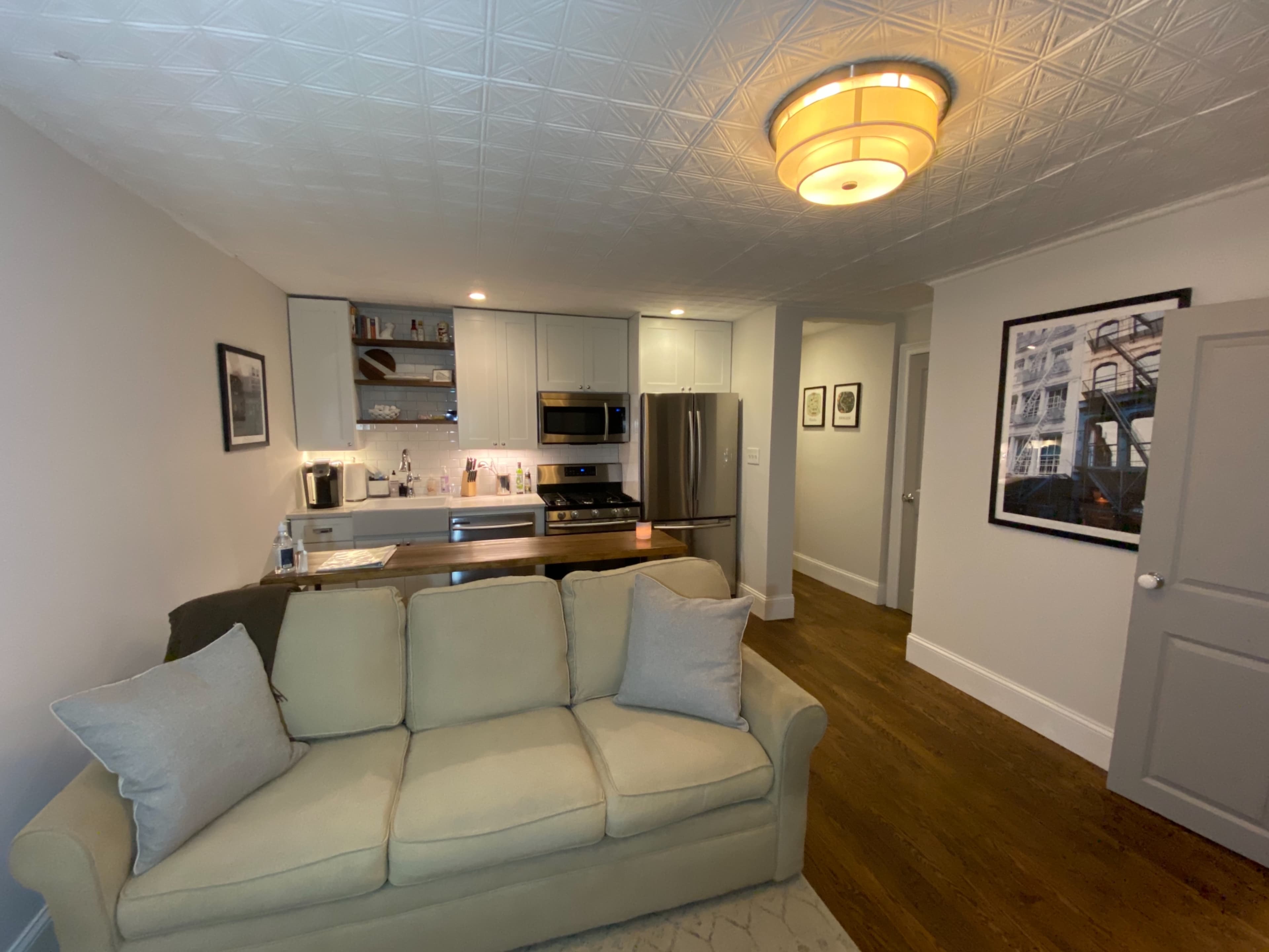 The image shows a modern living space featuring a beige sofa, a kitchen area with stainless steel appliances, and wood flooring.