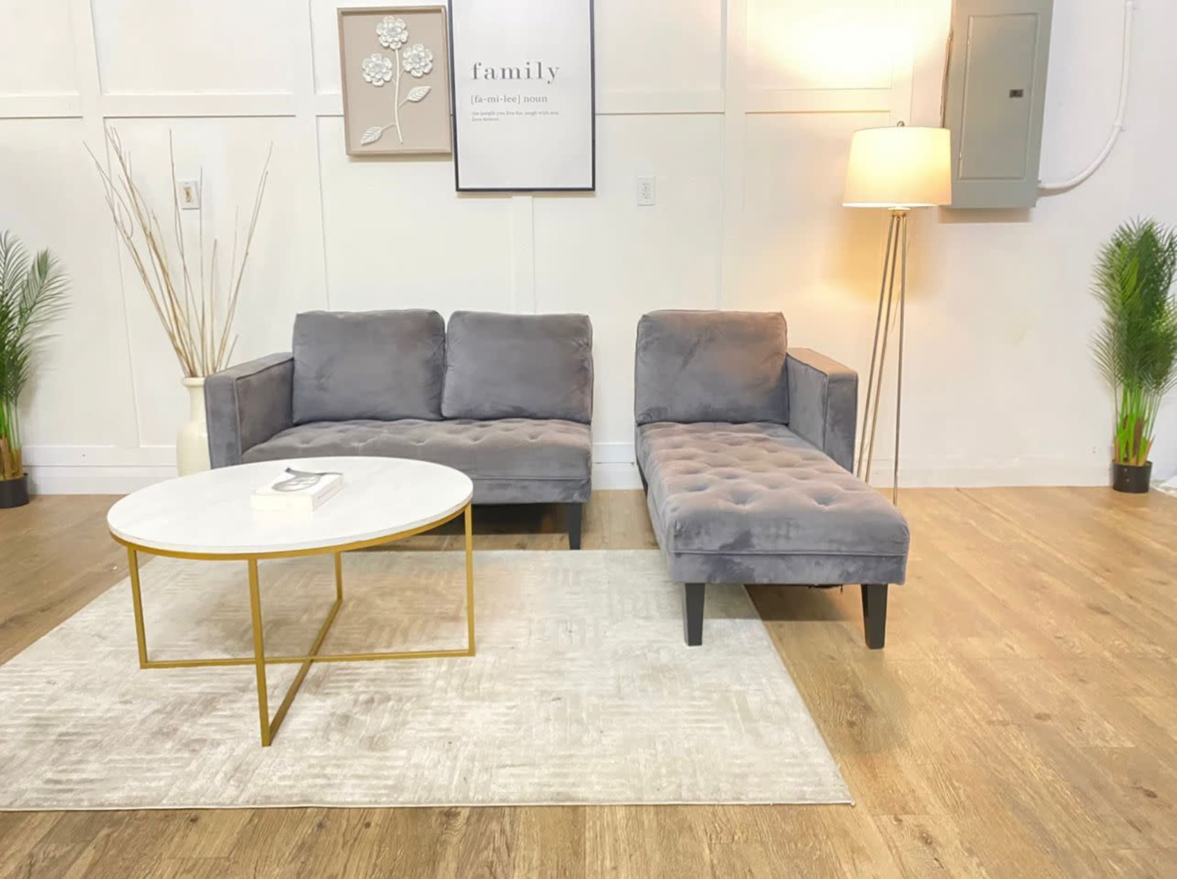 A modern living room setup with a gray couch, a chaise lounge, a round coffee table, and a lamp, set against a light-colored wall and wooden floor.