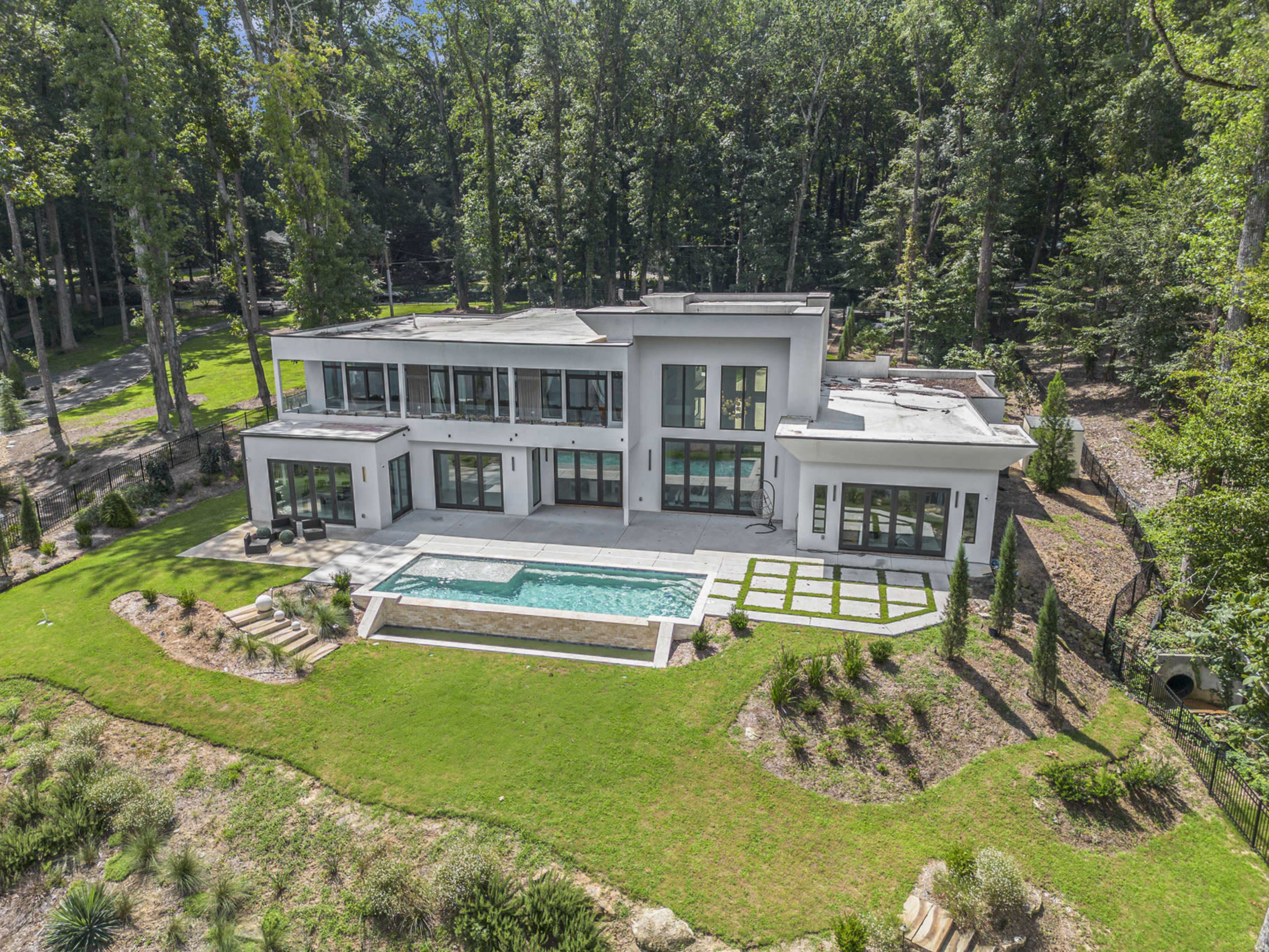 A modern two-story house with large windows, a swimming pool, and landscaped gardens is surrounded by dense trees.