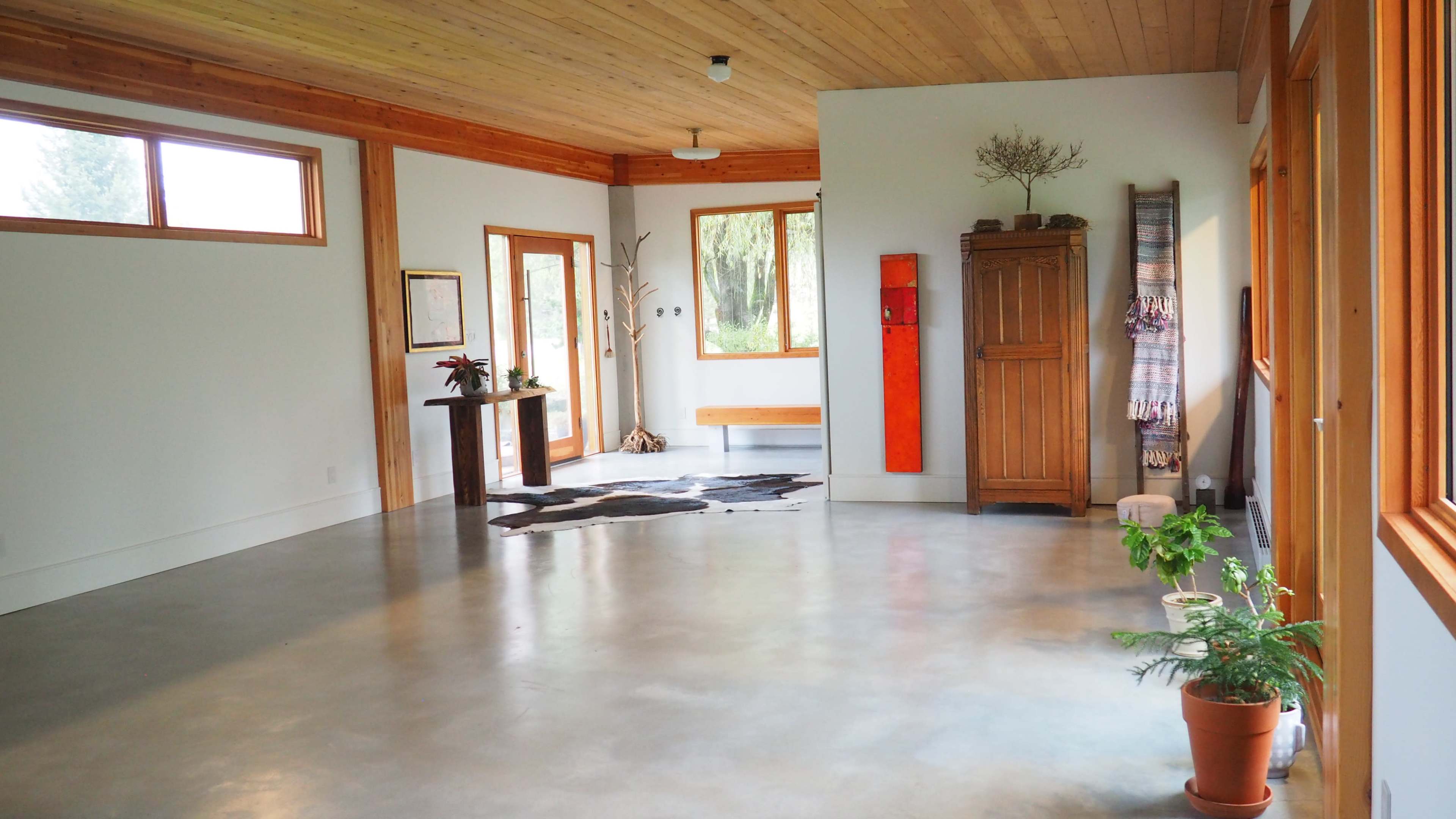 A spacious interior with a polished concrete floor, wooden beams, large windows, and minimal furnishings, including a cabinet and a small plant.