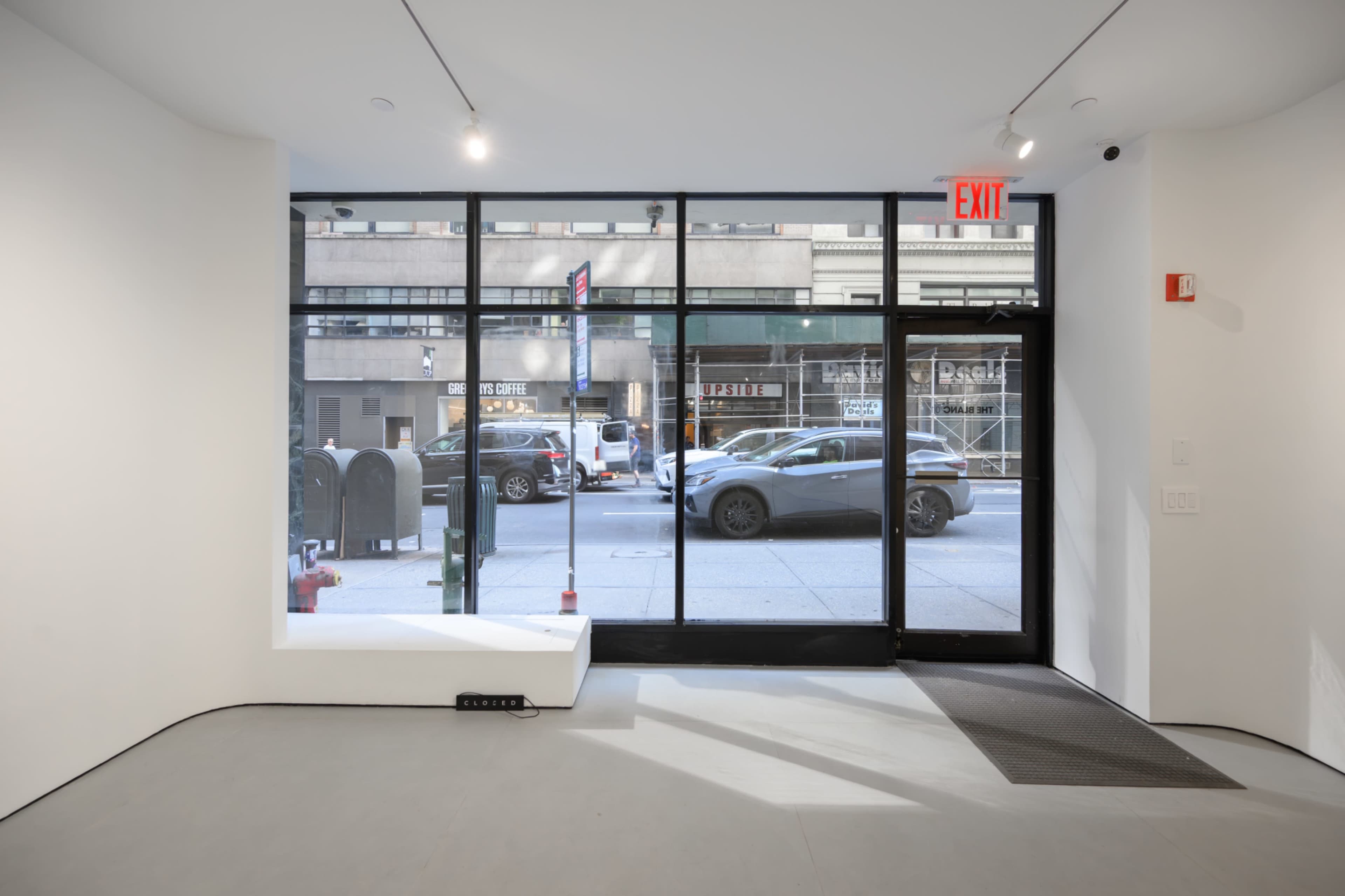 The image shows the interior of a minimalist storefront with large glass windows facing a city street.