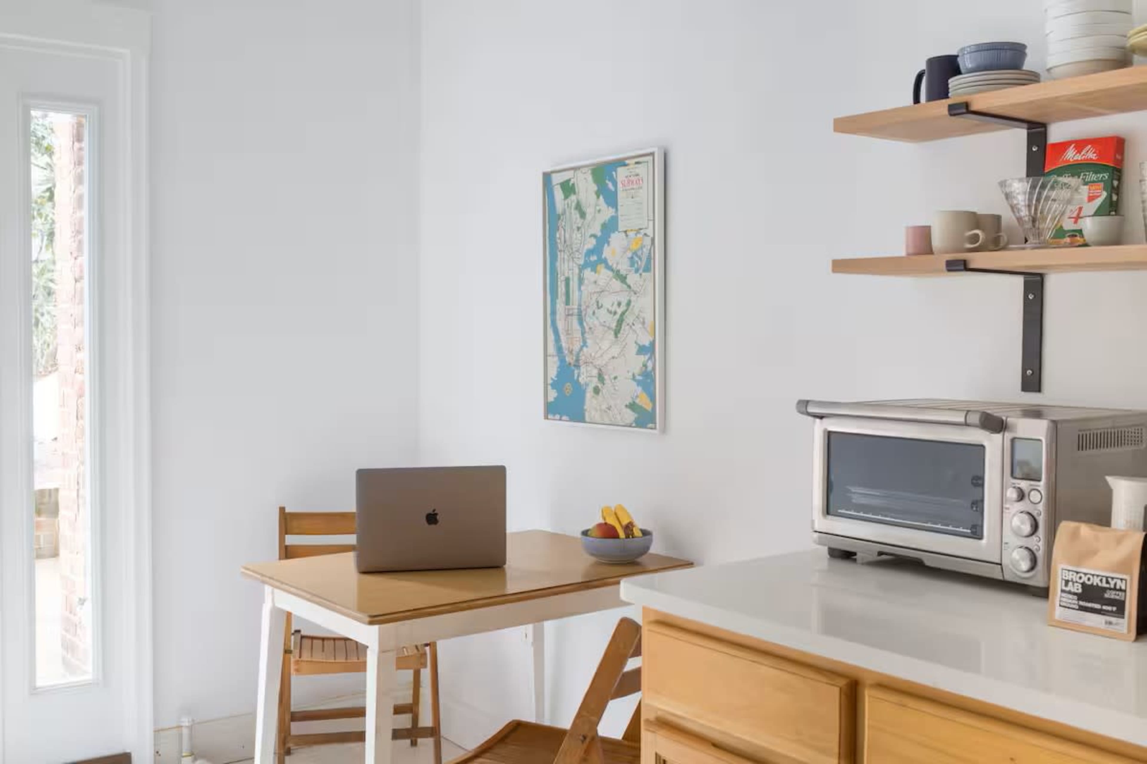A small kitchen features a wooden table with a laptop, a countertop toaster oven, and open shelving displaying various dishes and a map on the wall.