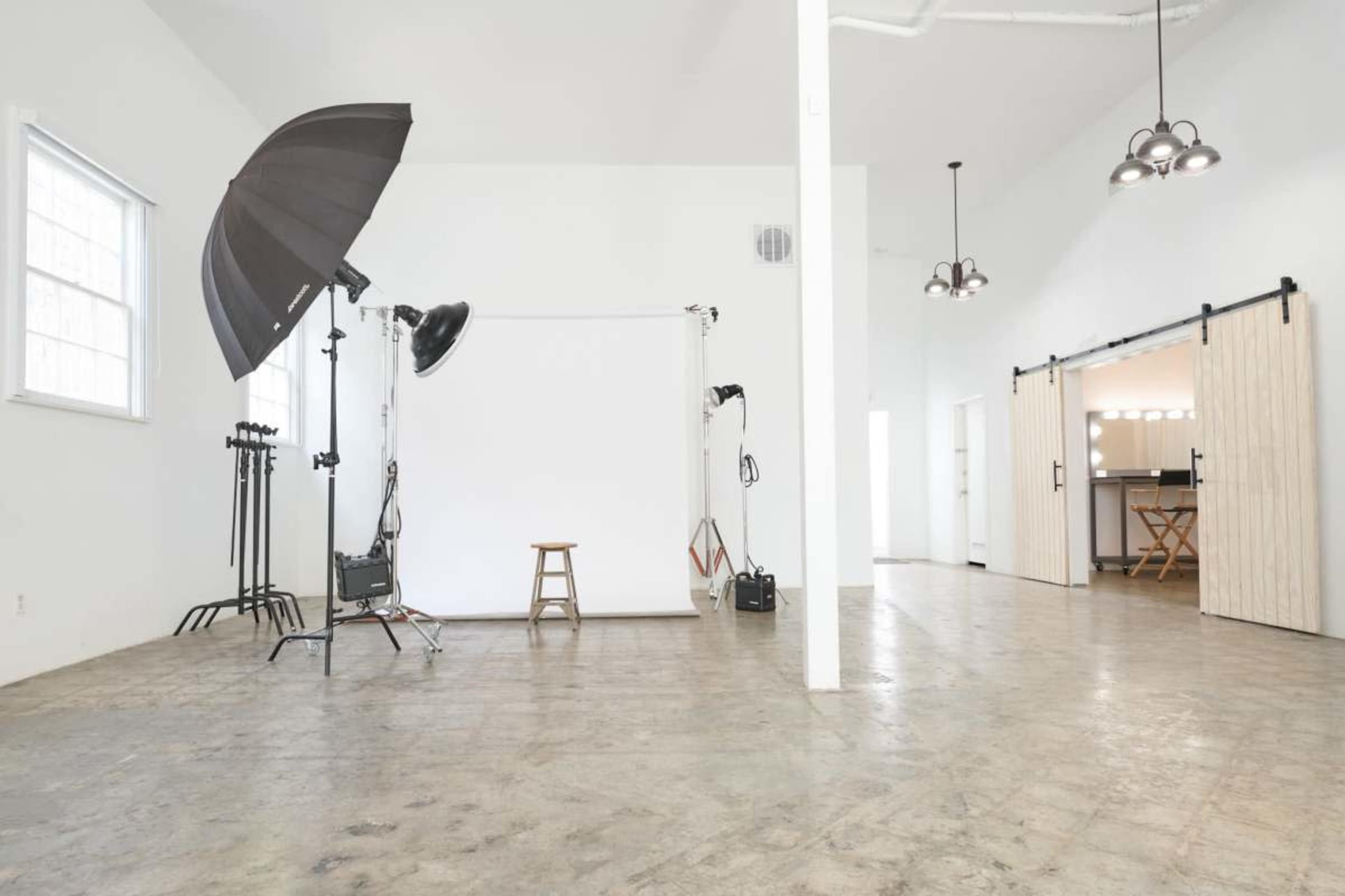 The image shows a spacious photography studio with a simple backdrop, lighting equipment, and a stool set against a light-colored wall.