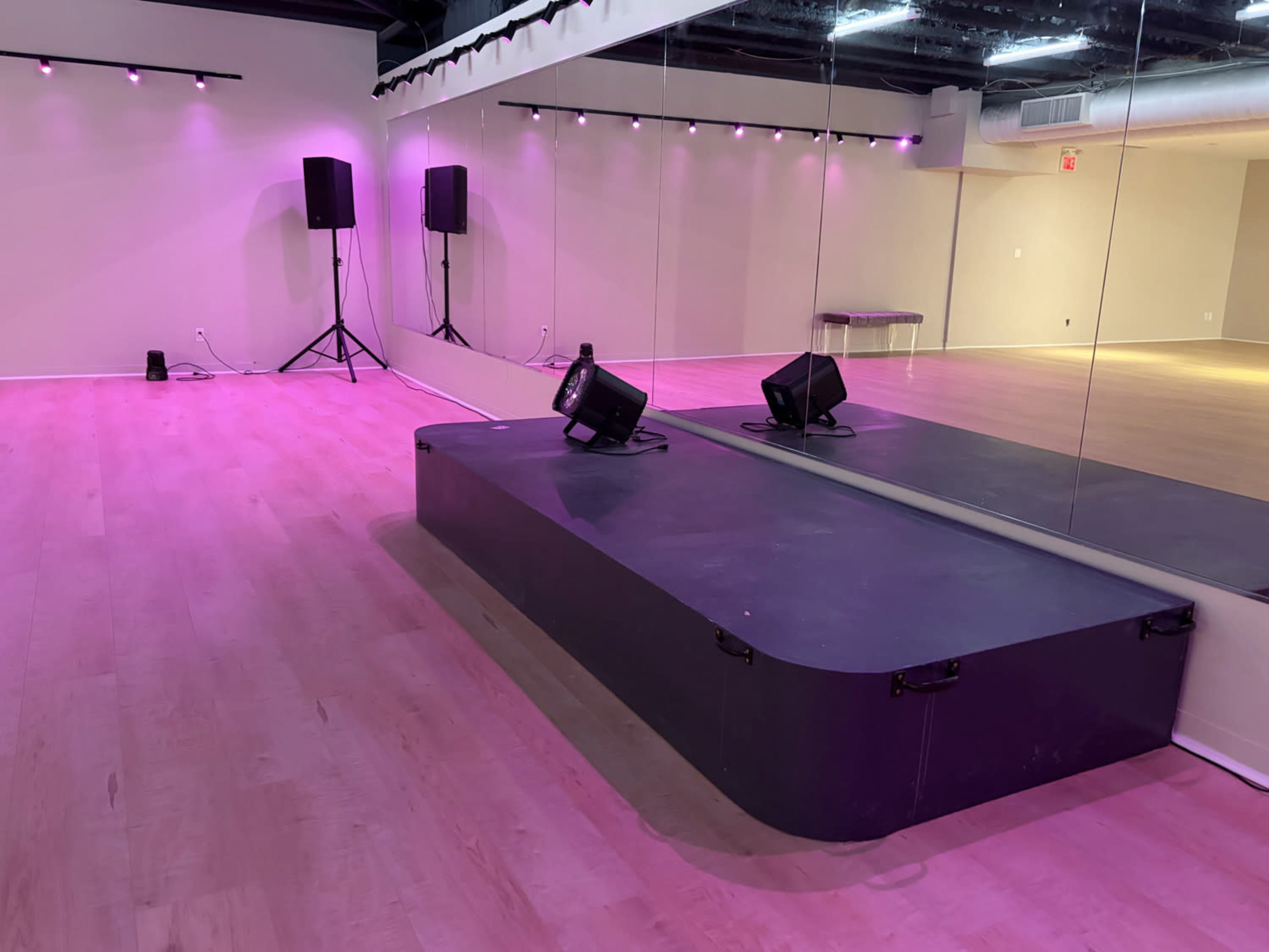 The image shows a dance studio with a mirrored wall, a raised platform in the center, and pink lighting along the ceiling.