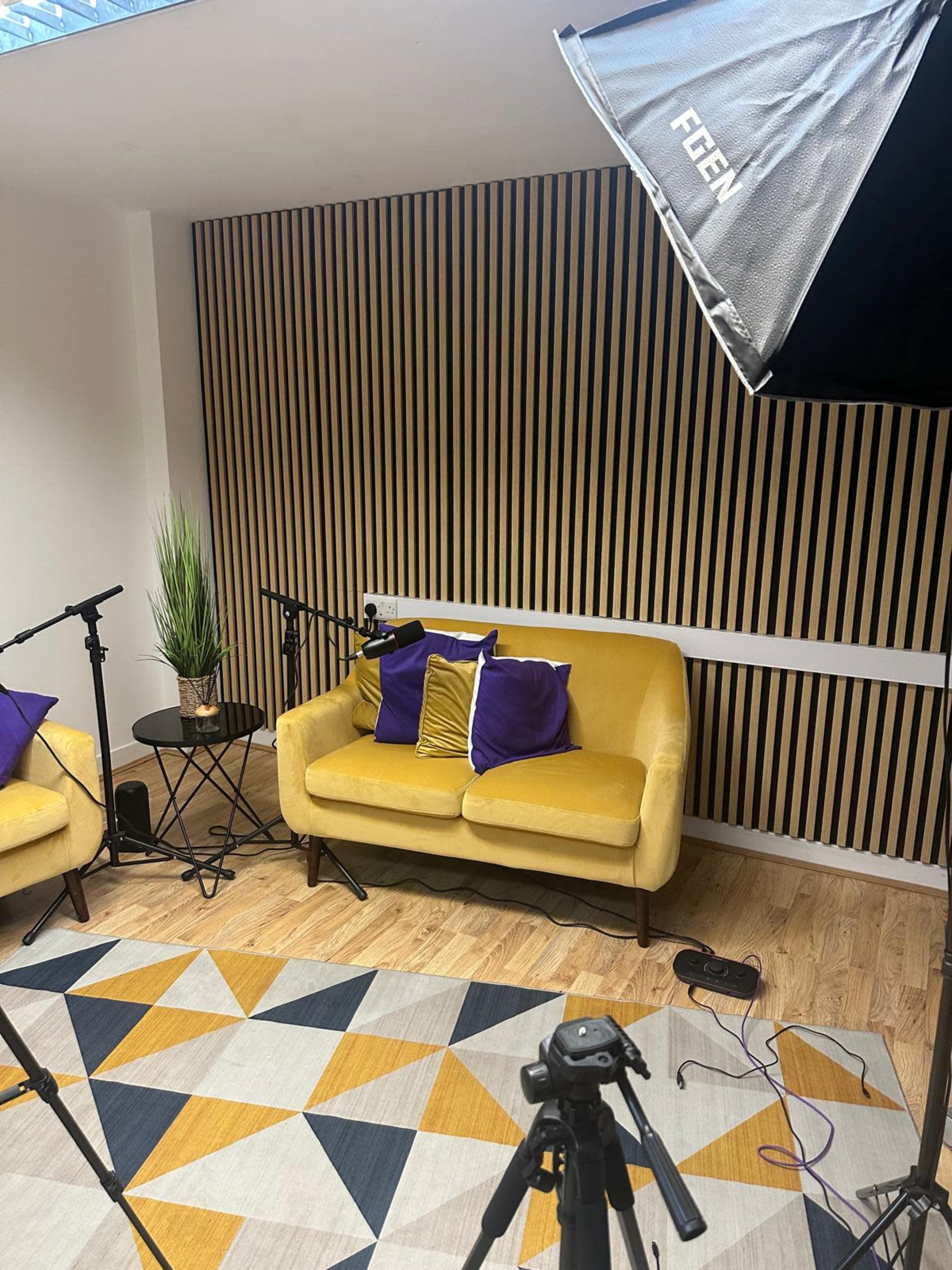 The image shows a modern indoor space featuring a yellow sofa with purple cushions, a geometric rug, a small round table, and studio lighting equipment.
