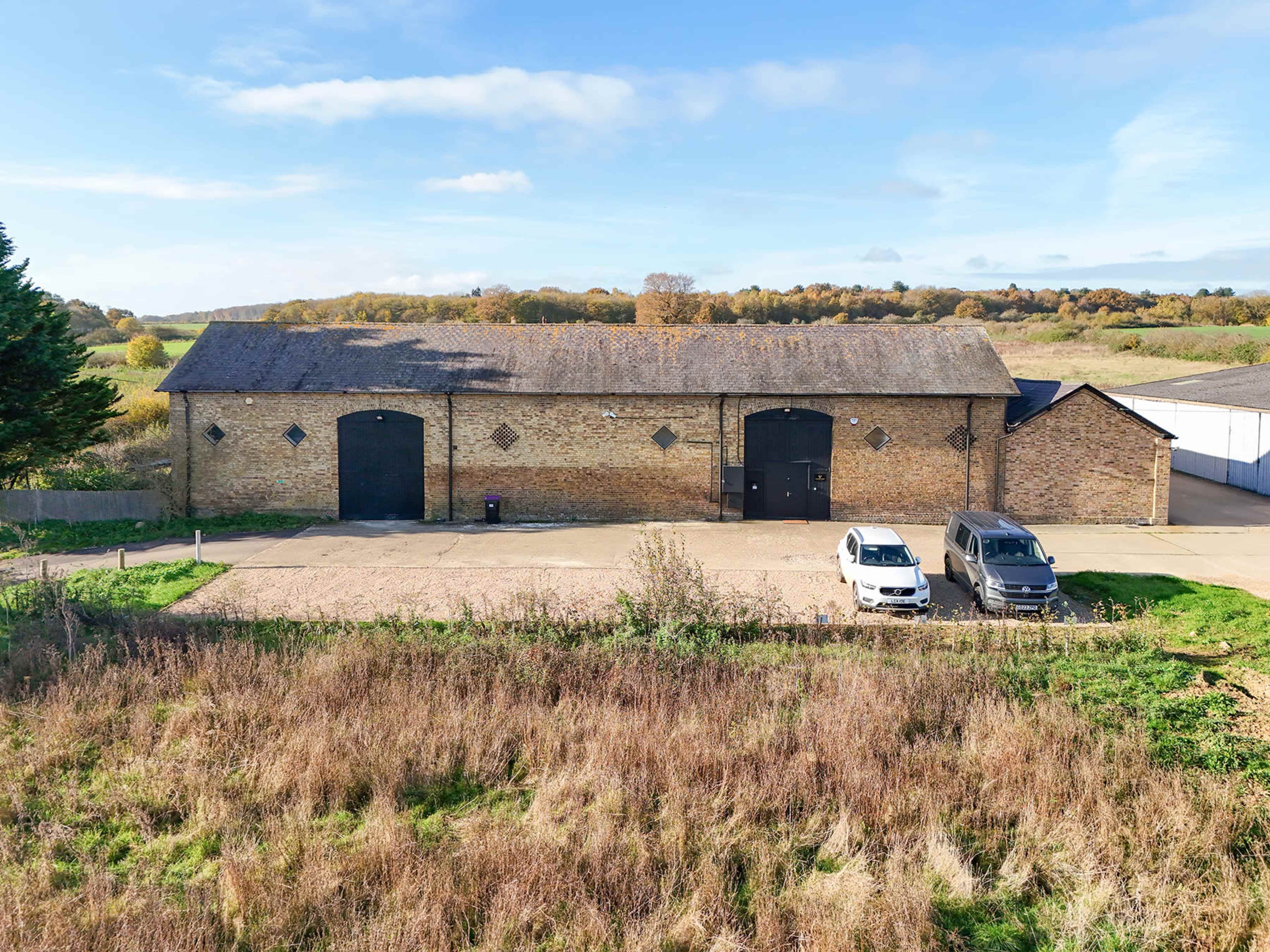 The image shows a large, brick warehouse with two vehicles parked in front and a grassy area surrounding it, set against a backdrop of trees and a clear sky.
