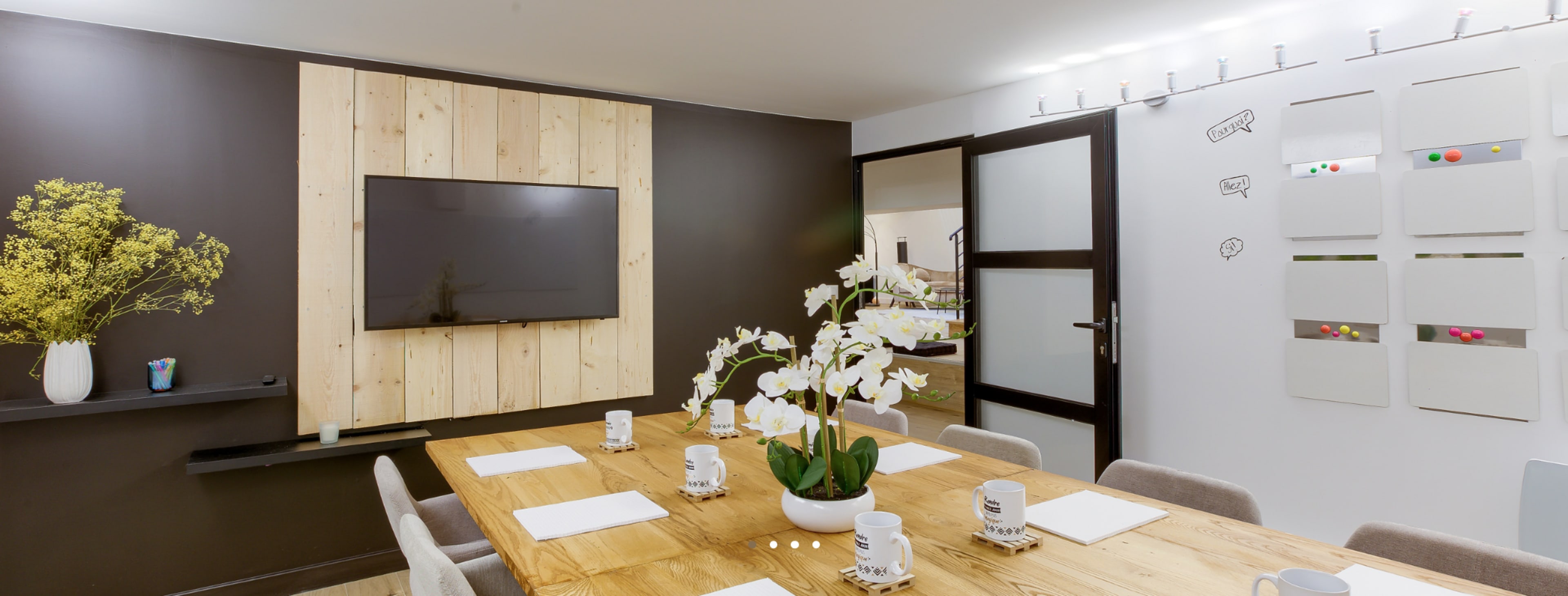 A modern meeting room with a wooden table, white orchids in a vase, a television mounted on a wooden panel, and a wall with whiteboards and colorful magnets.