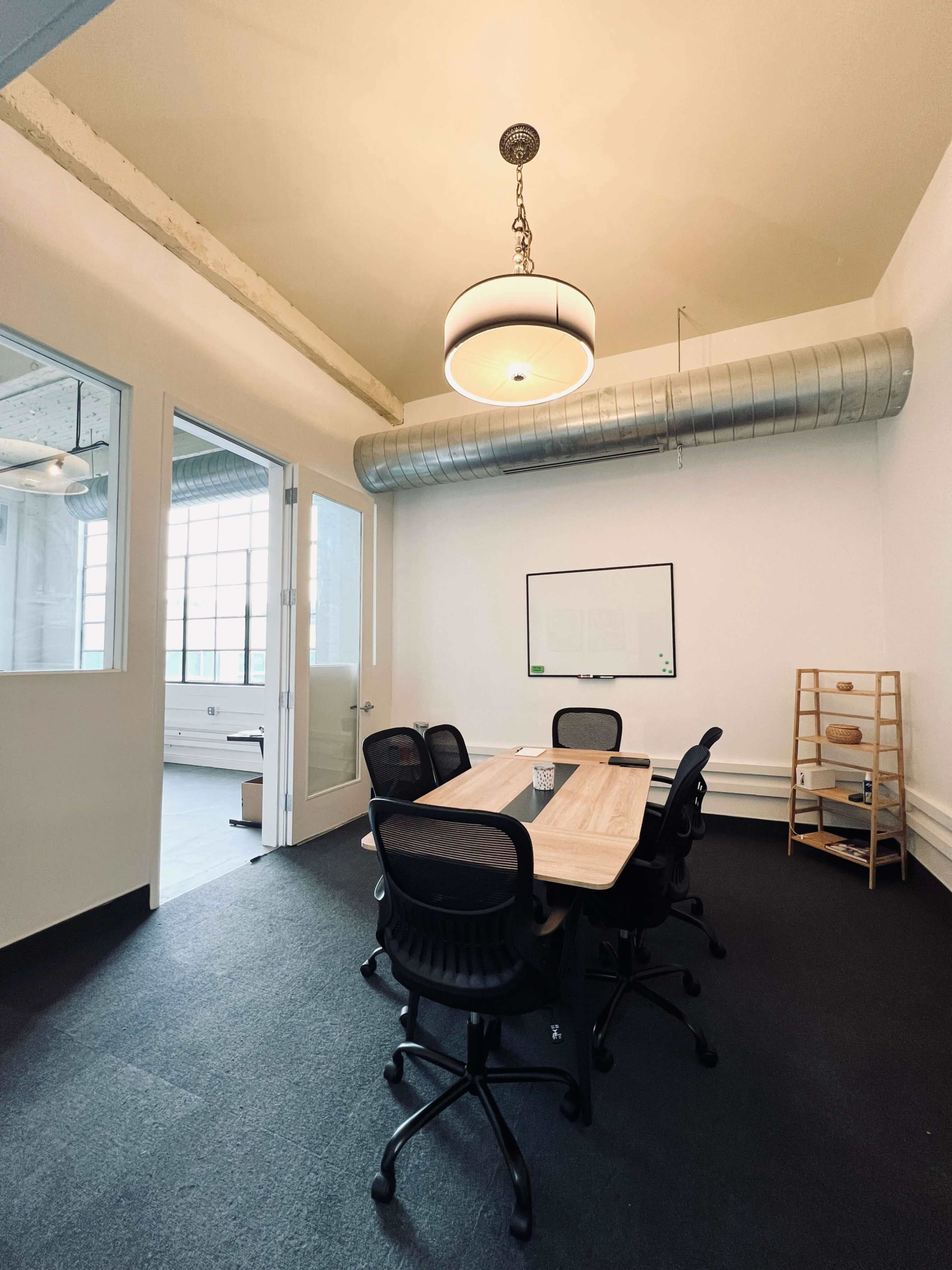 A conference room features a wooden table surrounded by black office chairs, with a large window and minimal decor.