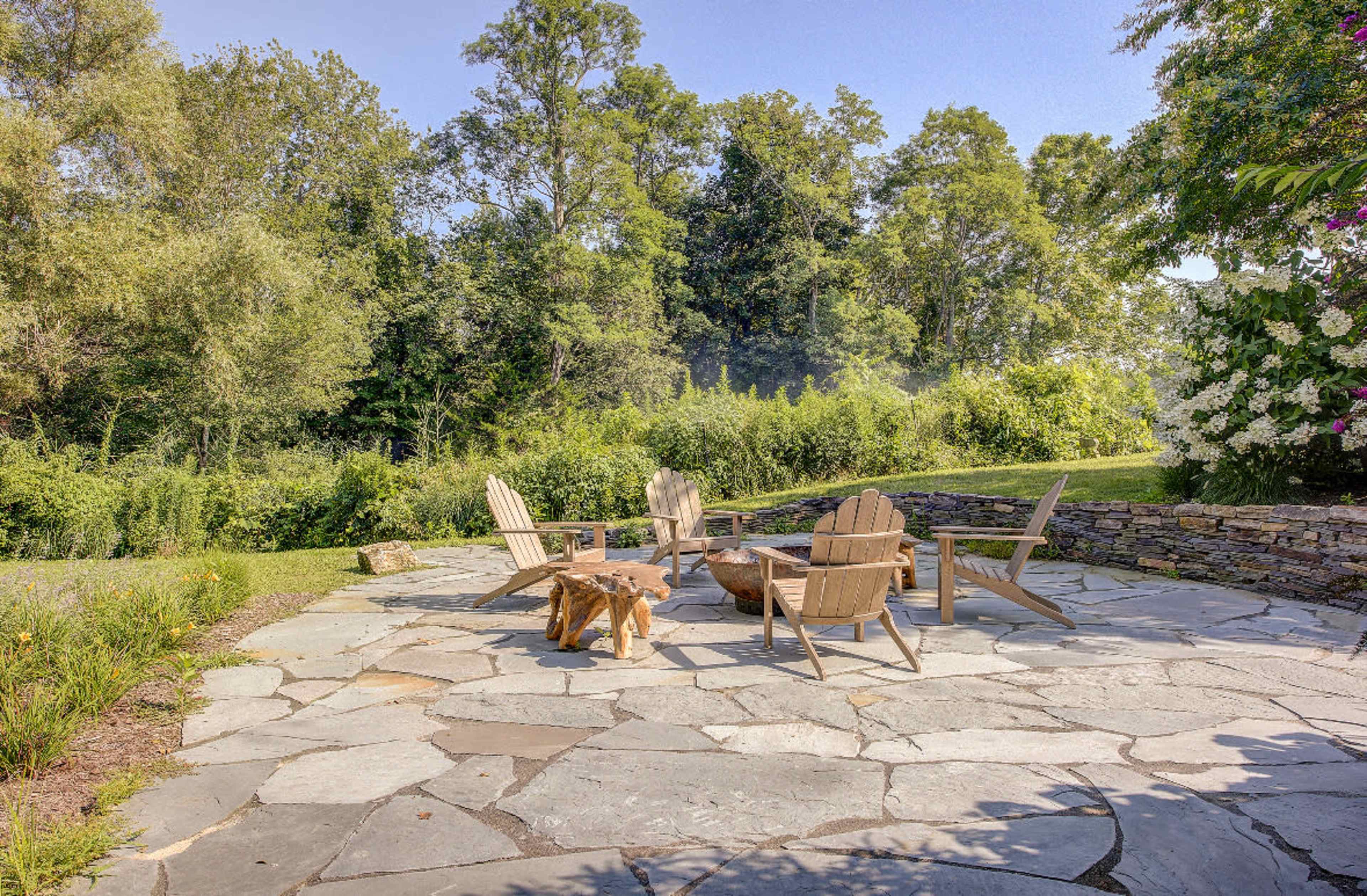 A stone patio features a circular arrangement of wooden Adirondack chairs around a fire pit, surrounded by greenery.