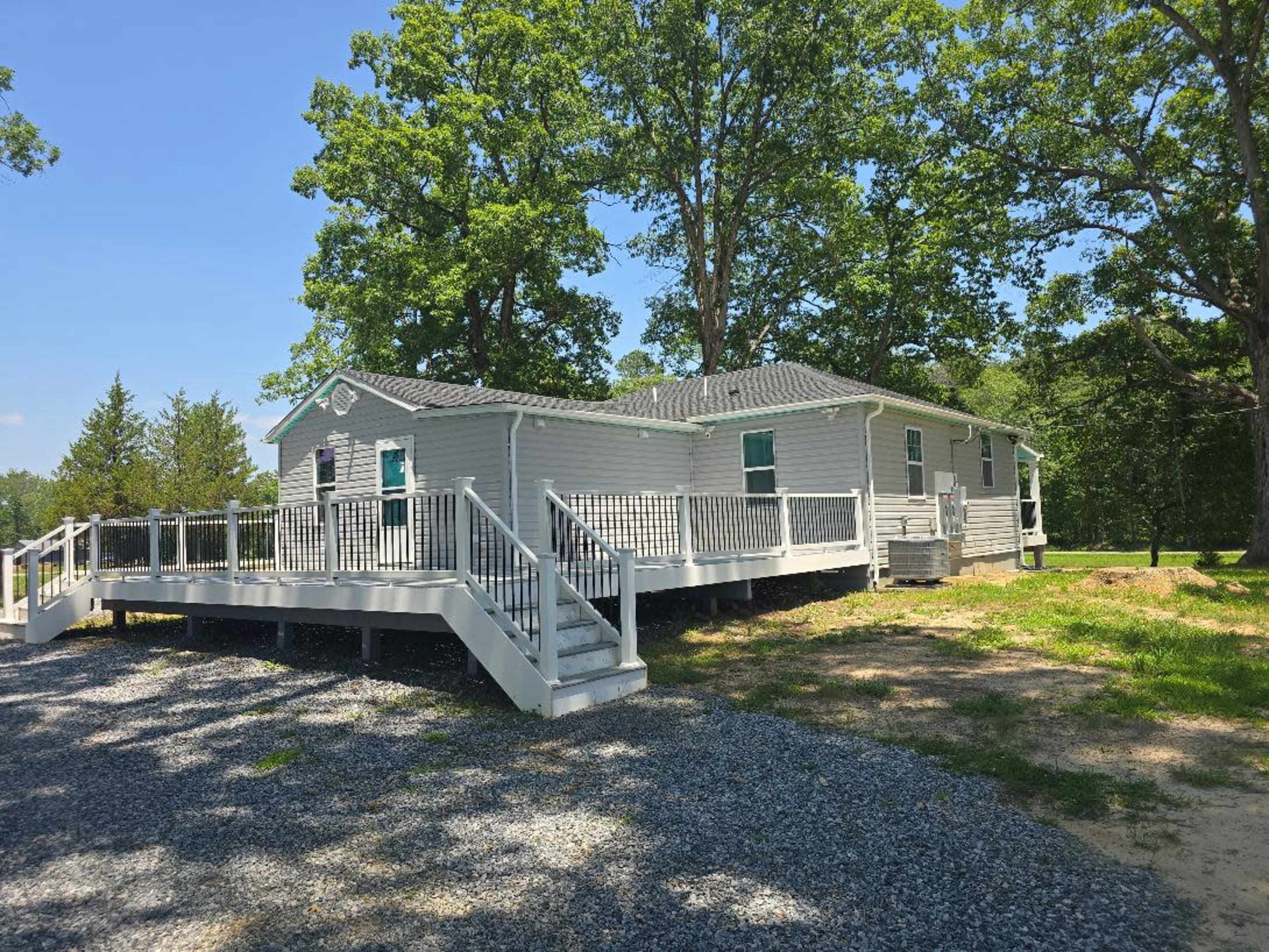 A gray house with a wraparound deck is surrounded by large trees and a gravel driveway.