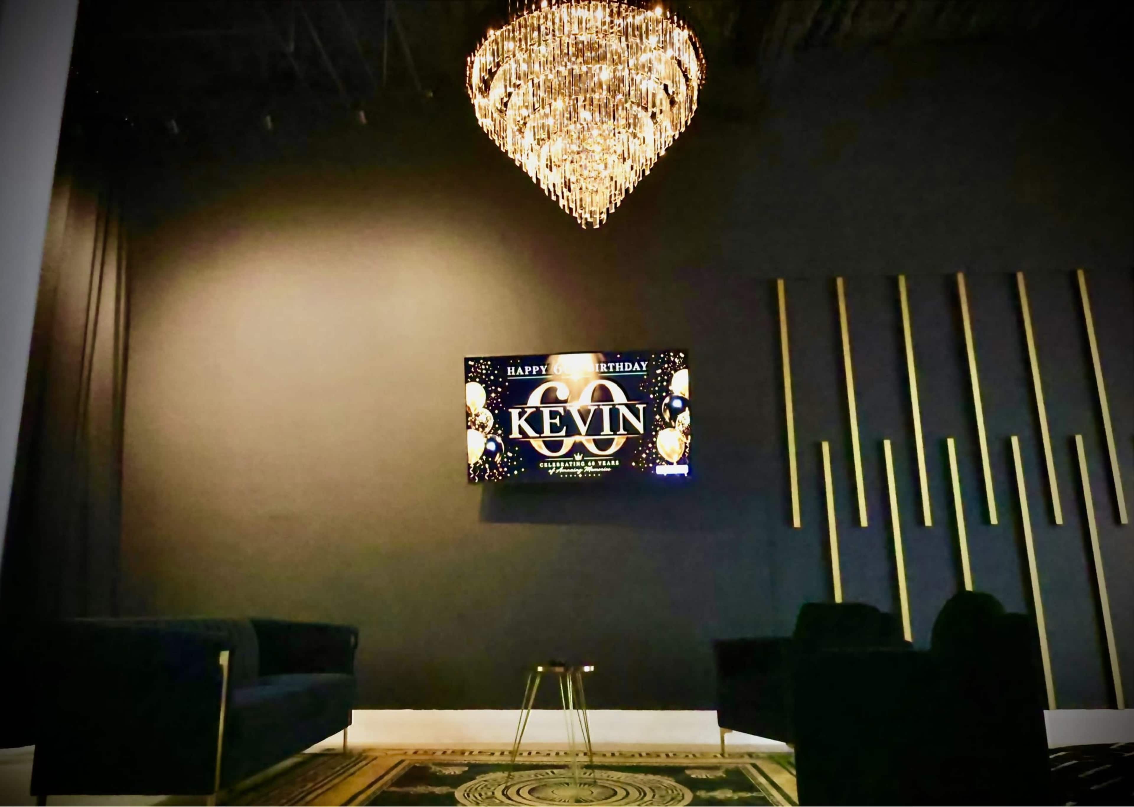 The image shows a modern event space featuring a large chandelier, a decorative wall with vertical slats, and a TV displaying a birthday message for "Kevin."
