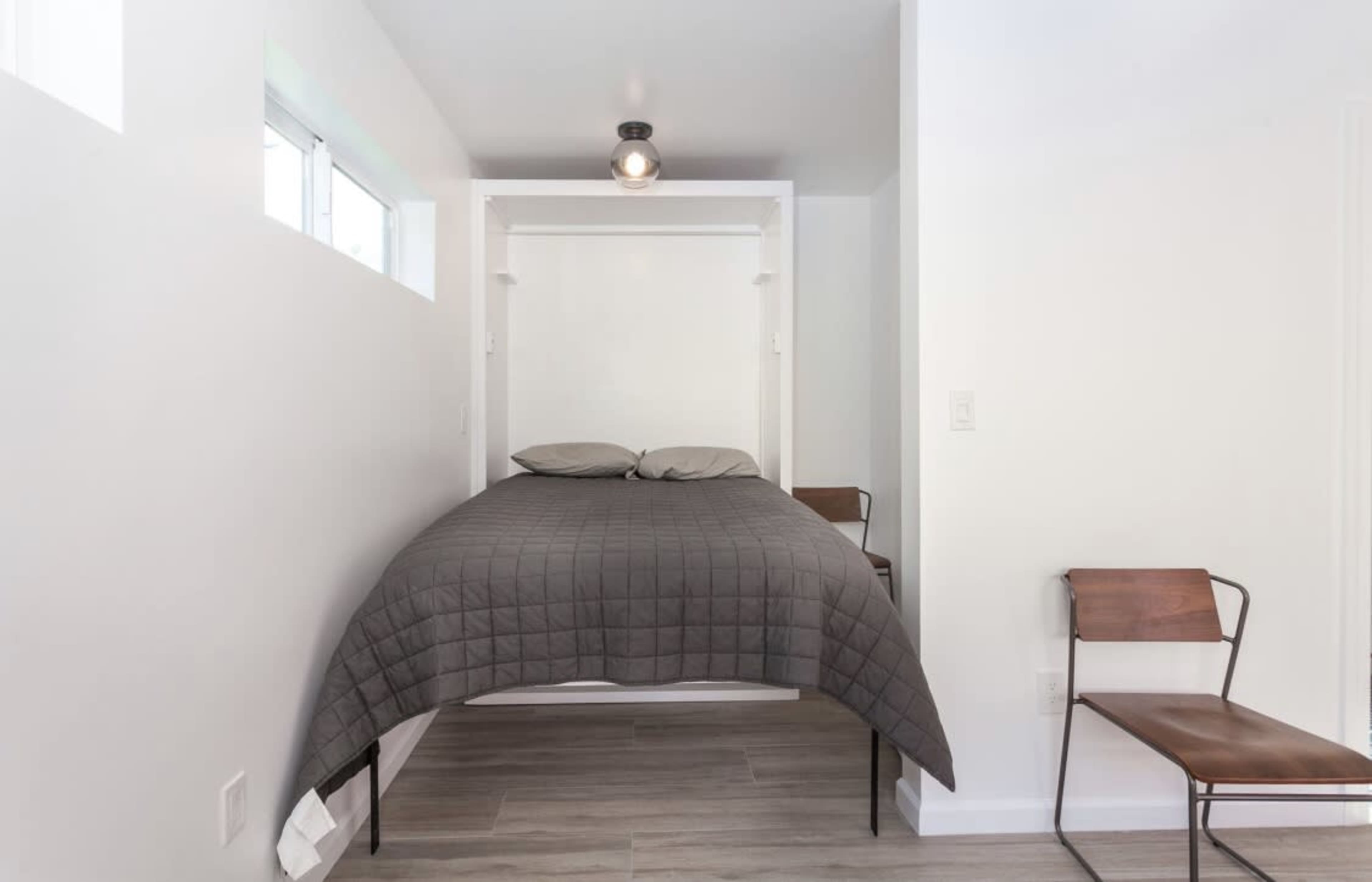 A small room featuring a wall-mounted bed with a gray quilt and two wooden chairs.