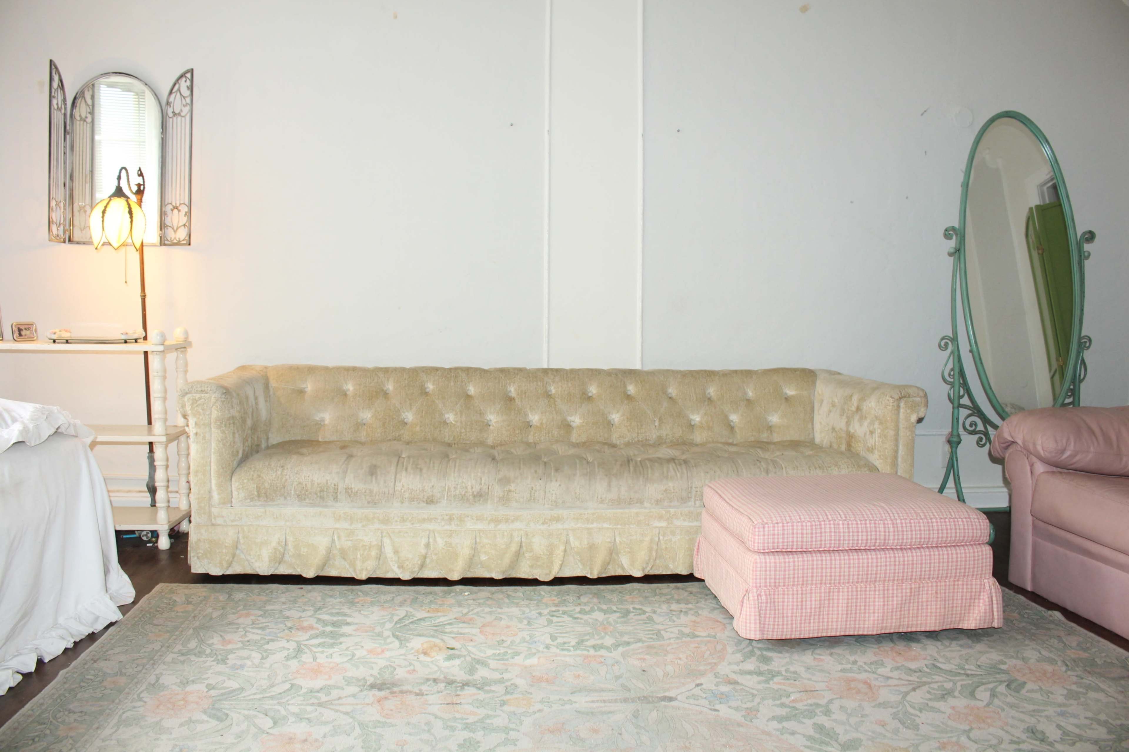 The scene features a vintage, tufted beige sofa beside a pink checkered ottoman, set against a plain white wall with a decorative mirror and lamp.