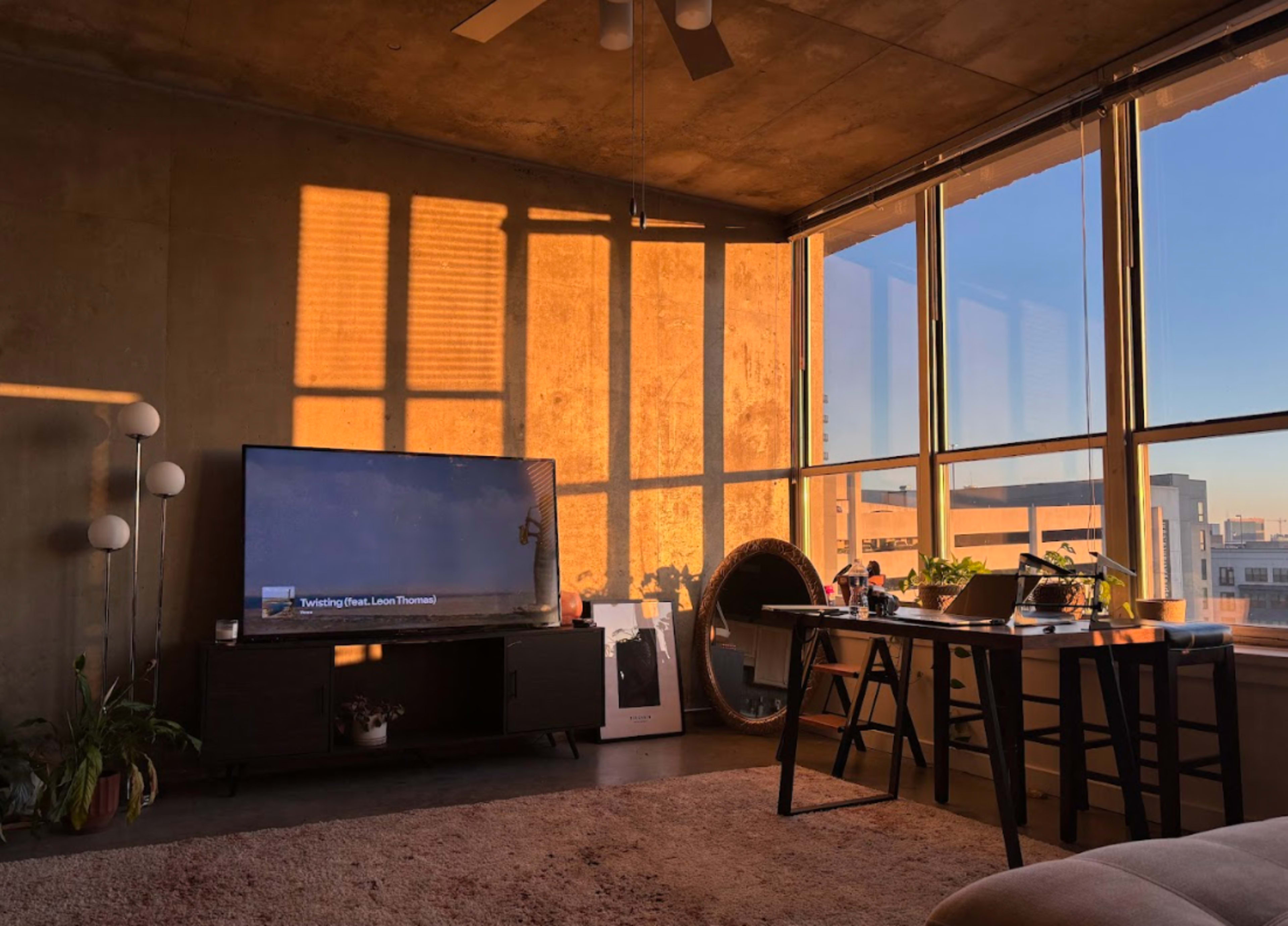The image shows a modern living room with a large window allowing sunlight to cast shadows on the walls, a television unit, a round mirror, and a small table with plants.