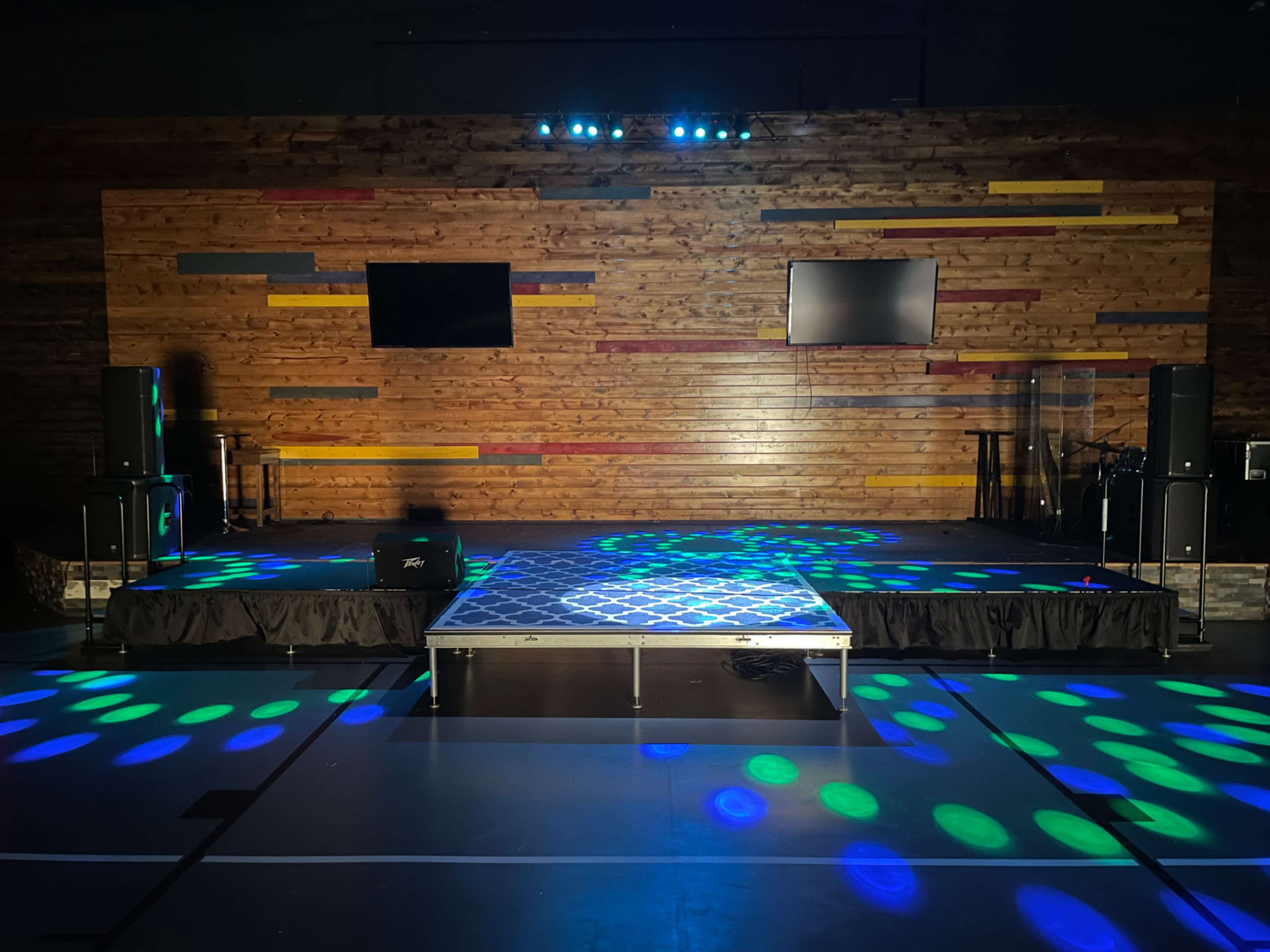 The image shows a stage with a wooden backdrop, two television screens, and colorful spotlights casting designs on the floor.