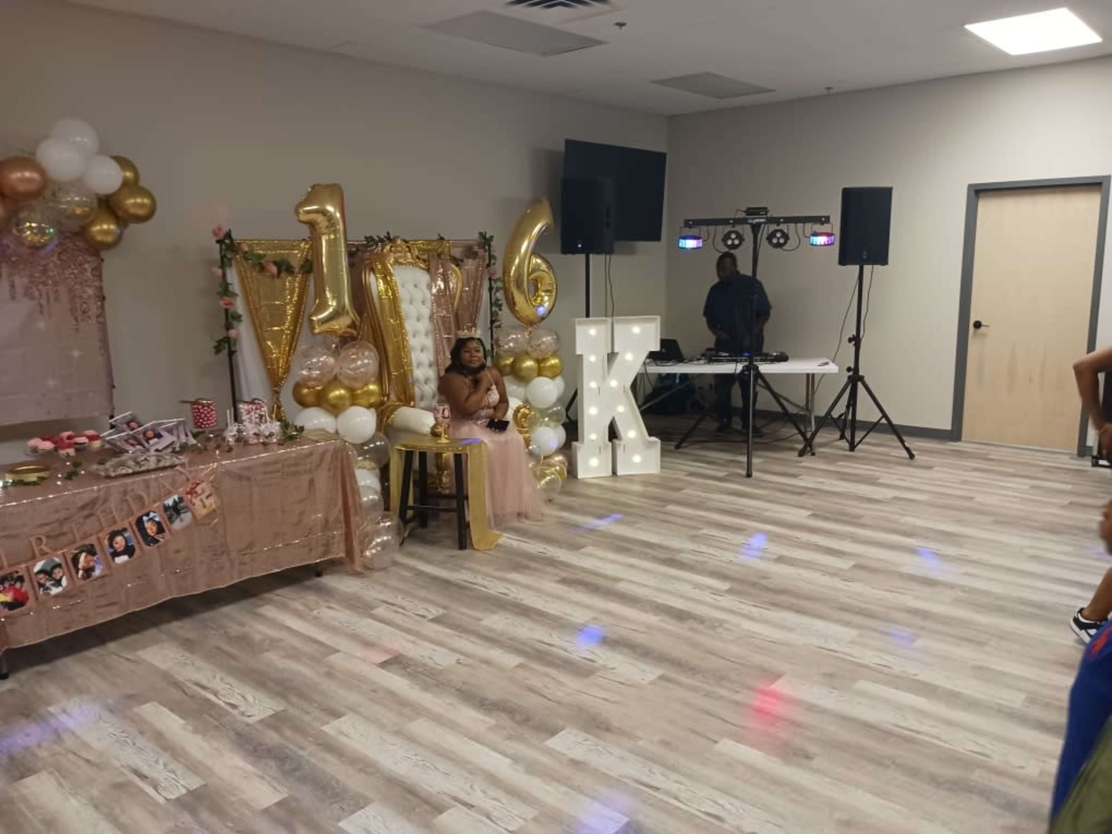A child sits on a chair in front of a decorated table with a "16" sign and party supplies in a reception hall.