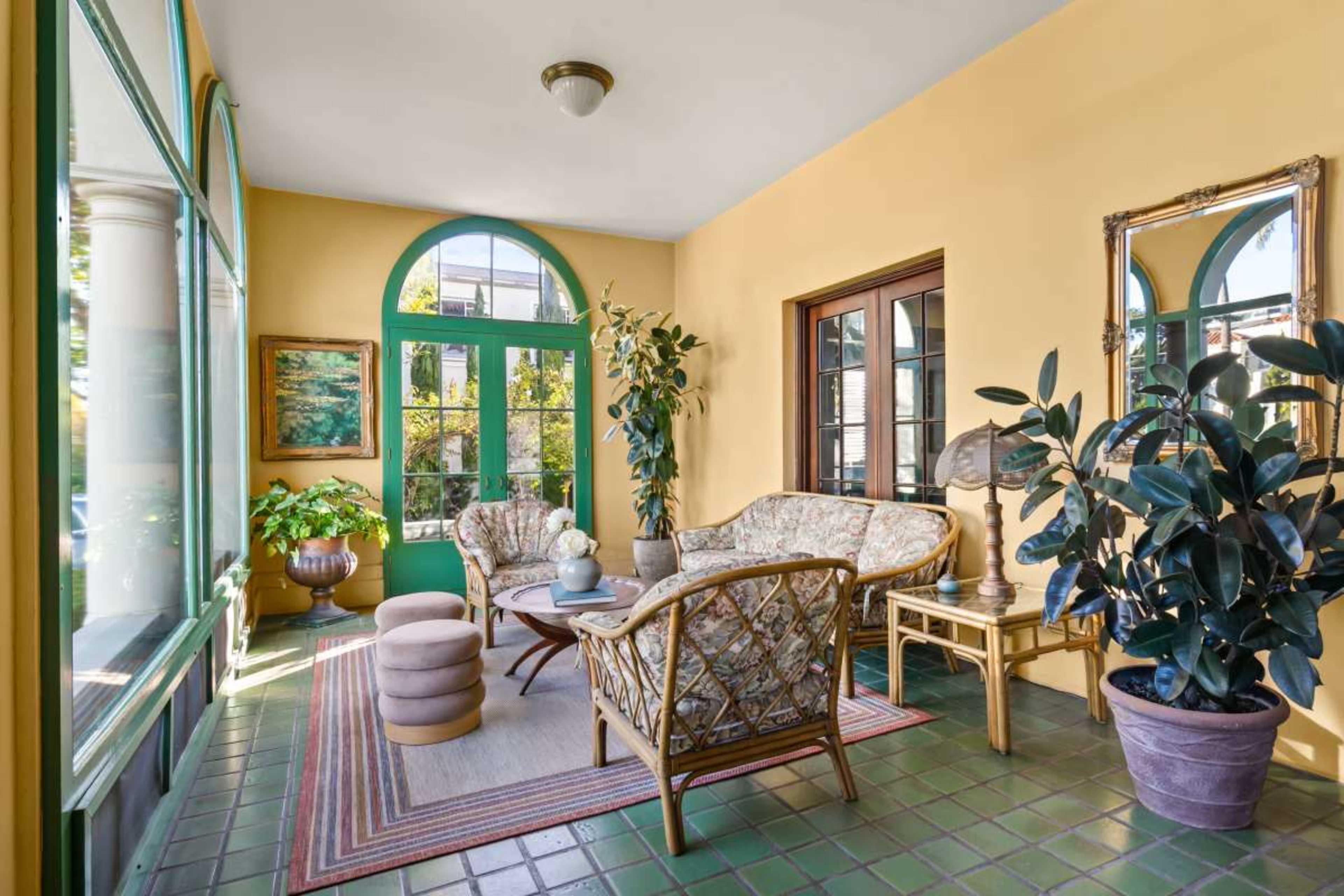 The image shows a sunlit sunroom with a floral-patterned seating arrangement, a small round table, and potted plants, set against yellow walls and green trim.