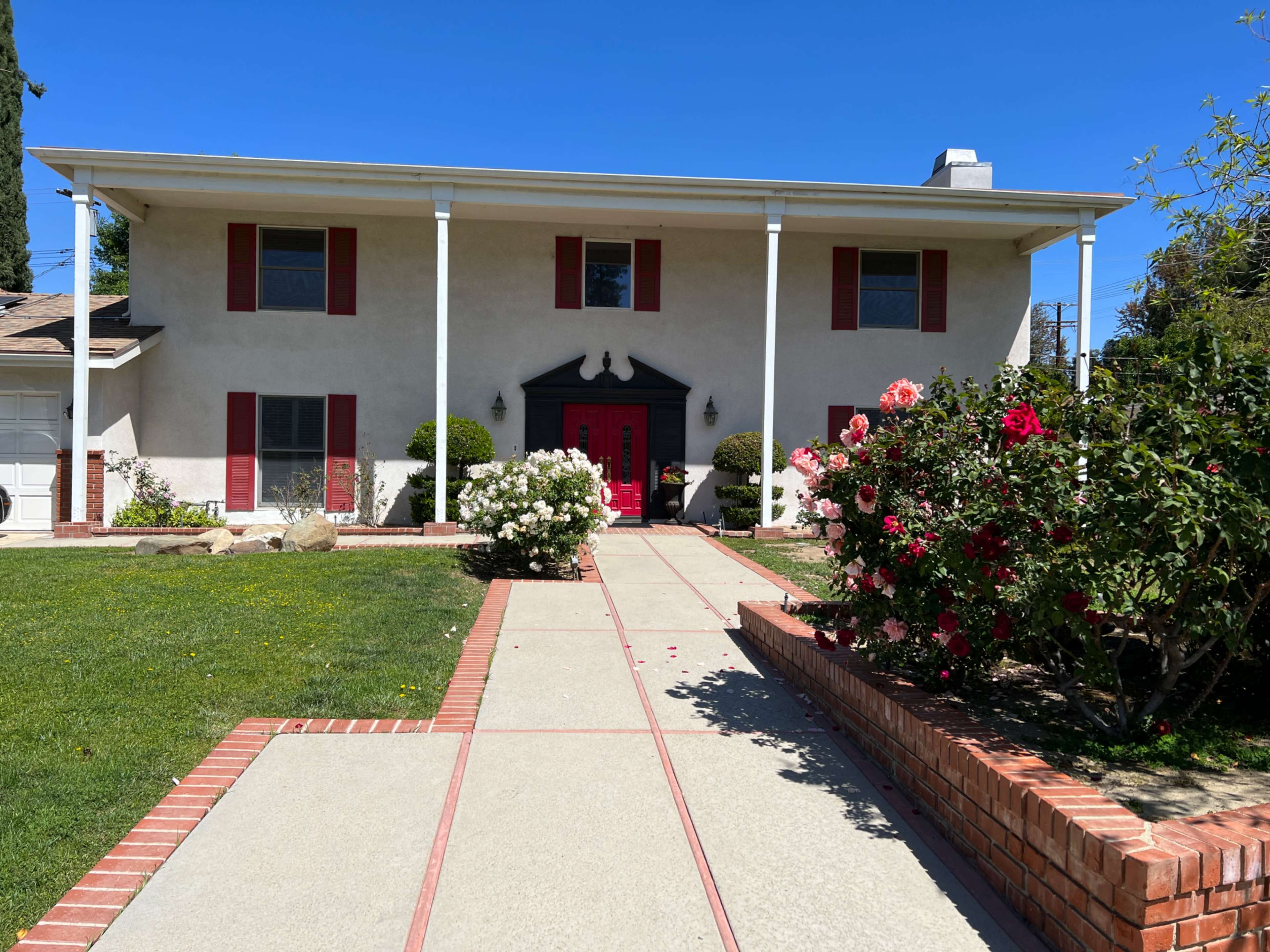 A two-story house with red shutters and a red front door, surrounded by flowering shrubs and a concrete pathway.