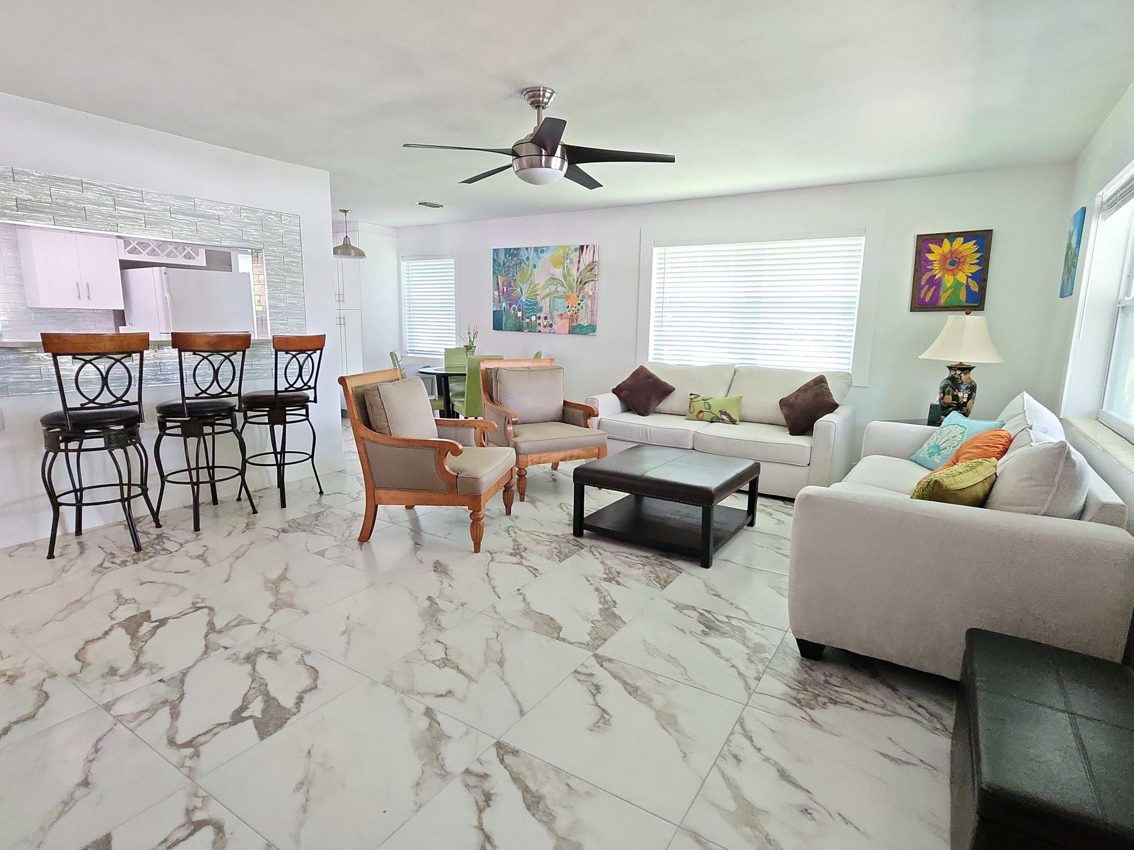 A modern living room with white furniture, marble-patterned flooring, and large windows providing natural light.