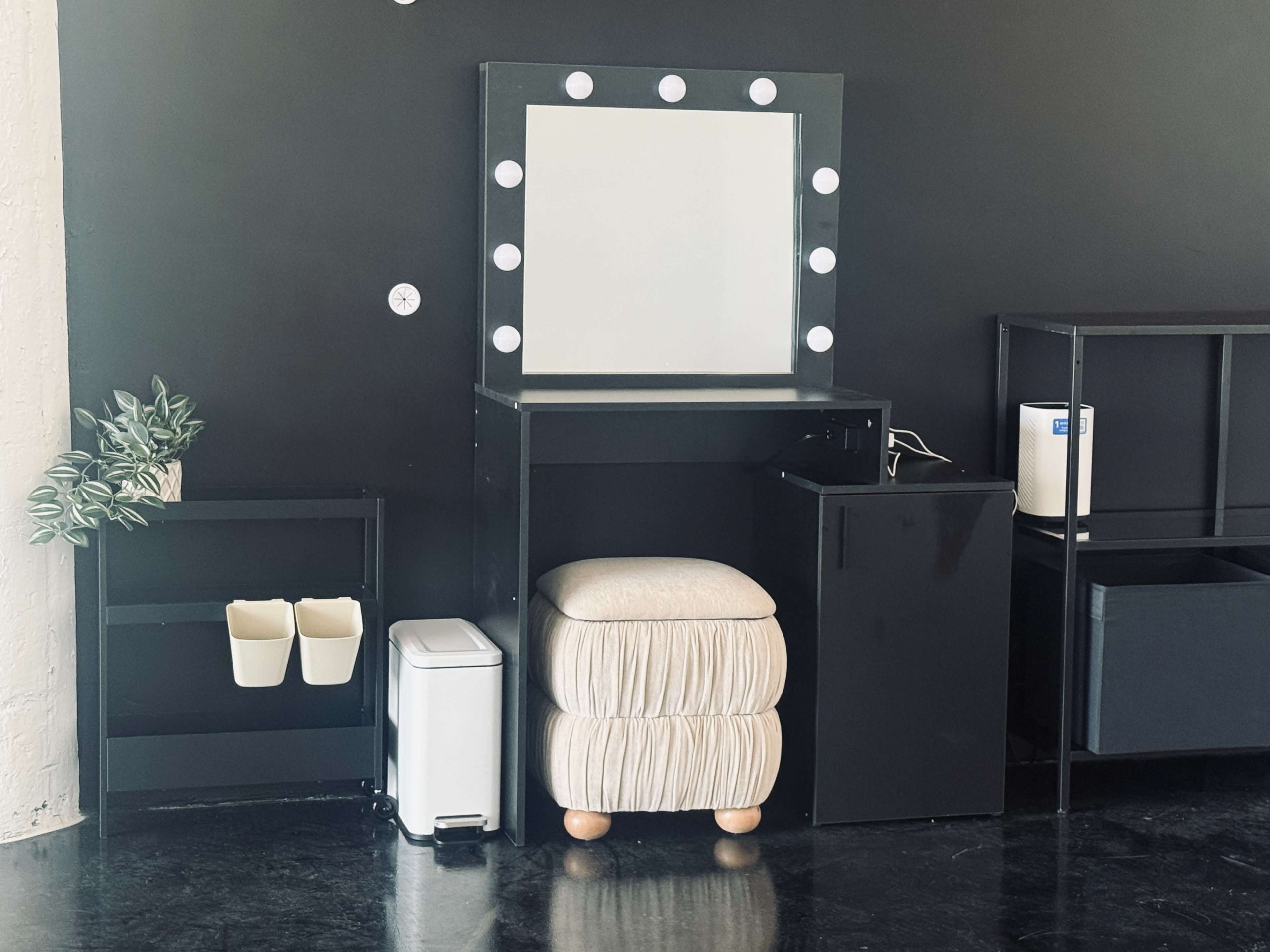 The image shows a minimalist vanity setup with a large mirror framed by lights, a stool, a small trash can, and a couple of shelves against a black wall.