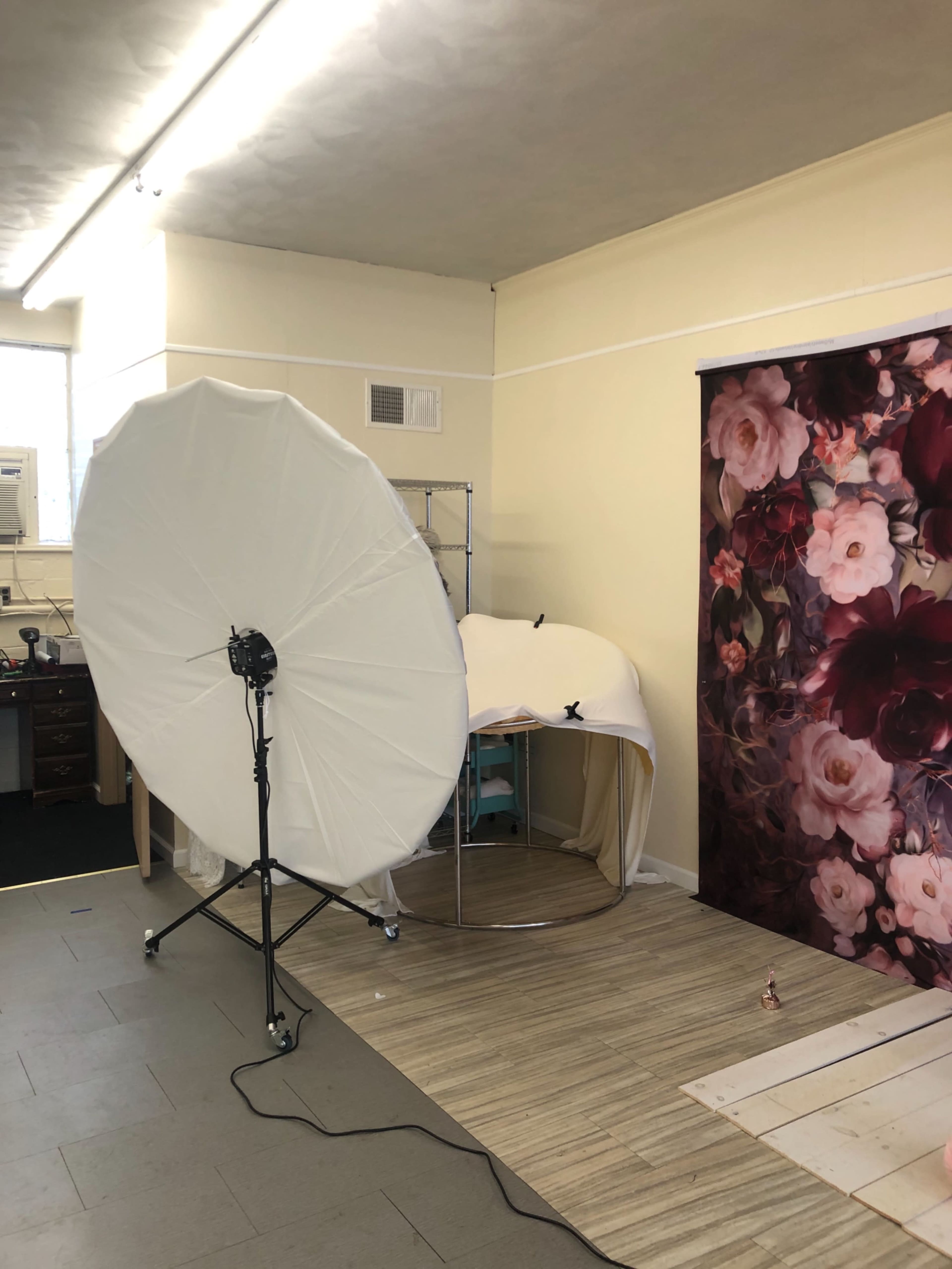 A photography studio setup featuring a large reflective umbrella light, a curved display rack covered with fabric, and a floral backdrop.
