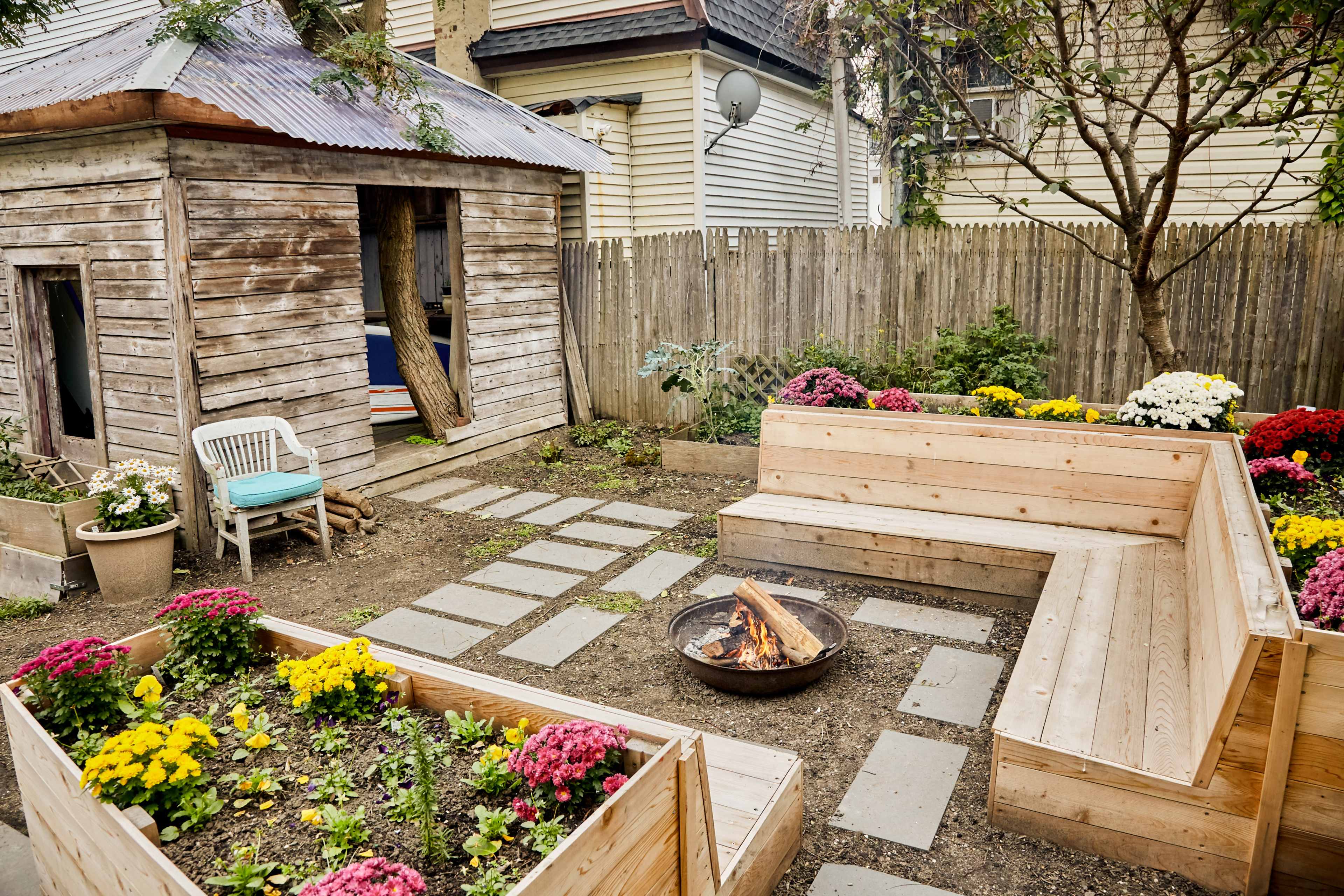 A cozy backyard with a wooden shed, a circular fire pit surrounded by wooden benches, and flower beds filled with colorful blooming plants.