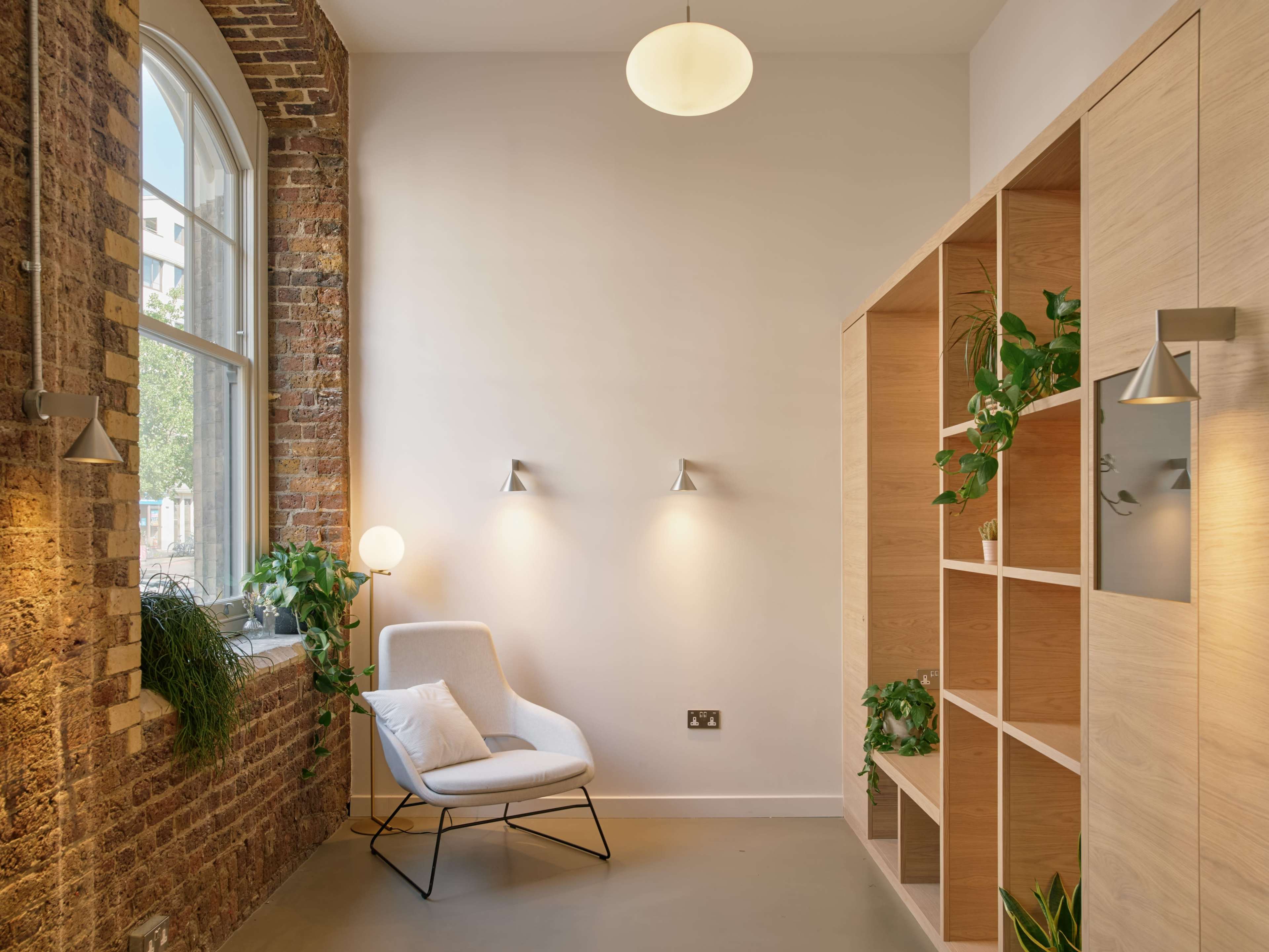 The image shows a modern interior featuring a white chair next to a wooden bookshelf adorned with plants, illuminated by wall sconces and a pendant light.