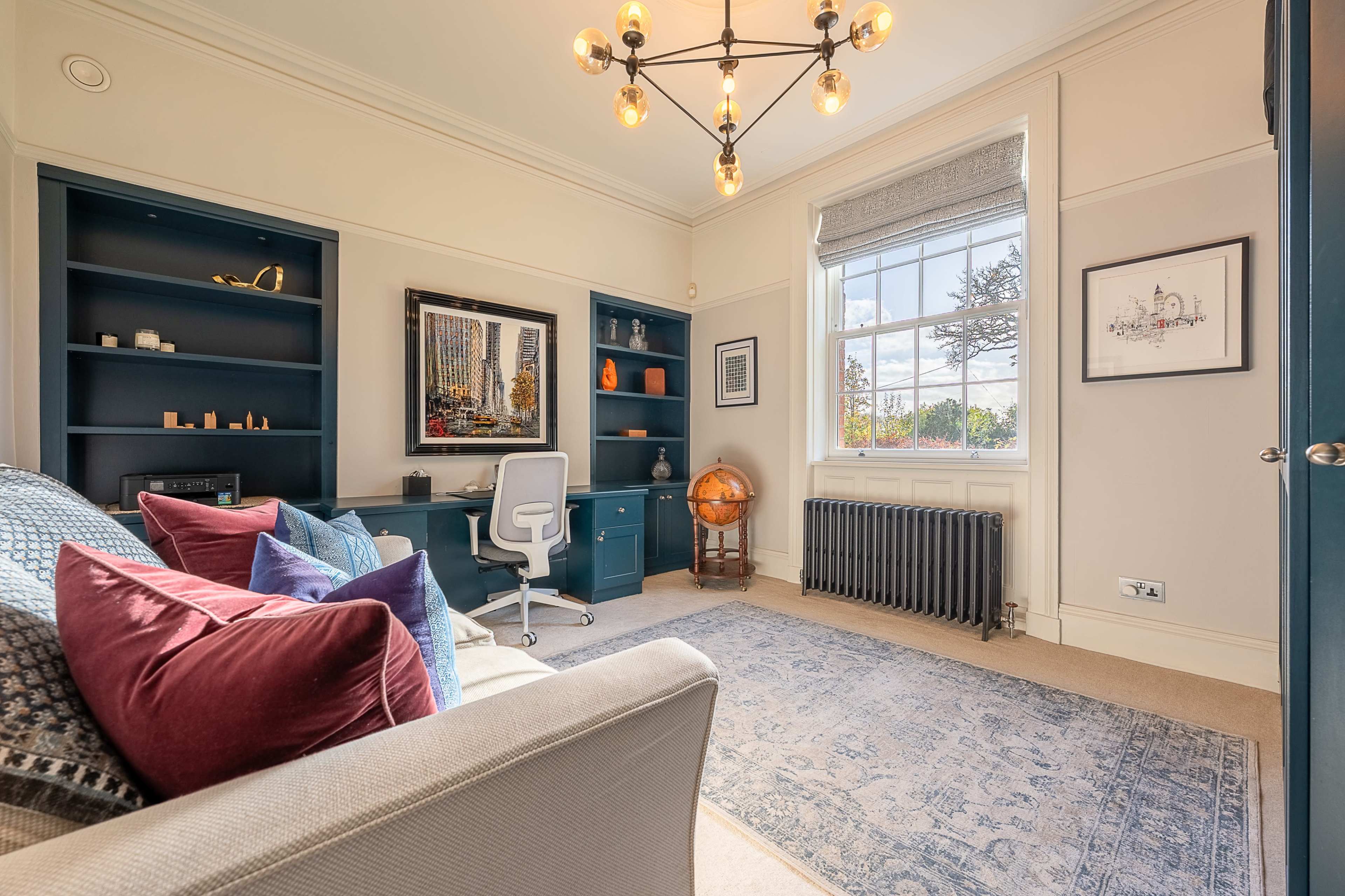 A well-furnished home office features a desk and chair, shelves with decorative items, and a large window providing natural light.